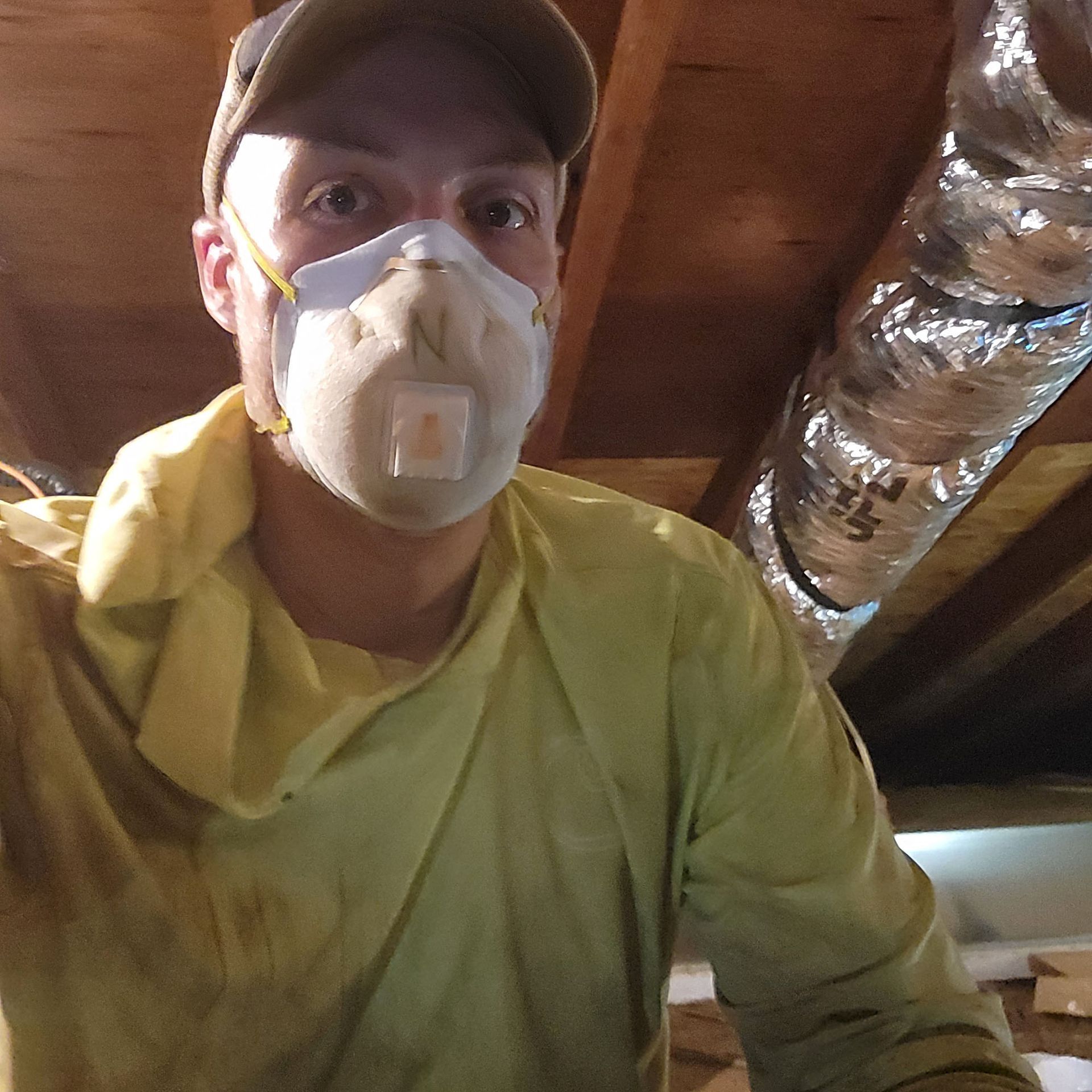 Person in a cap and mask, wearing a yellow shirt, working in an attic with exposed ductwork.