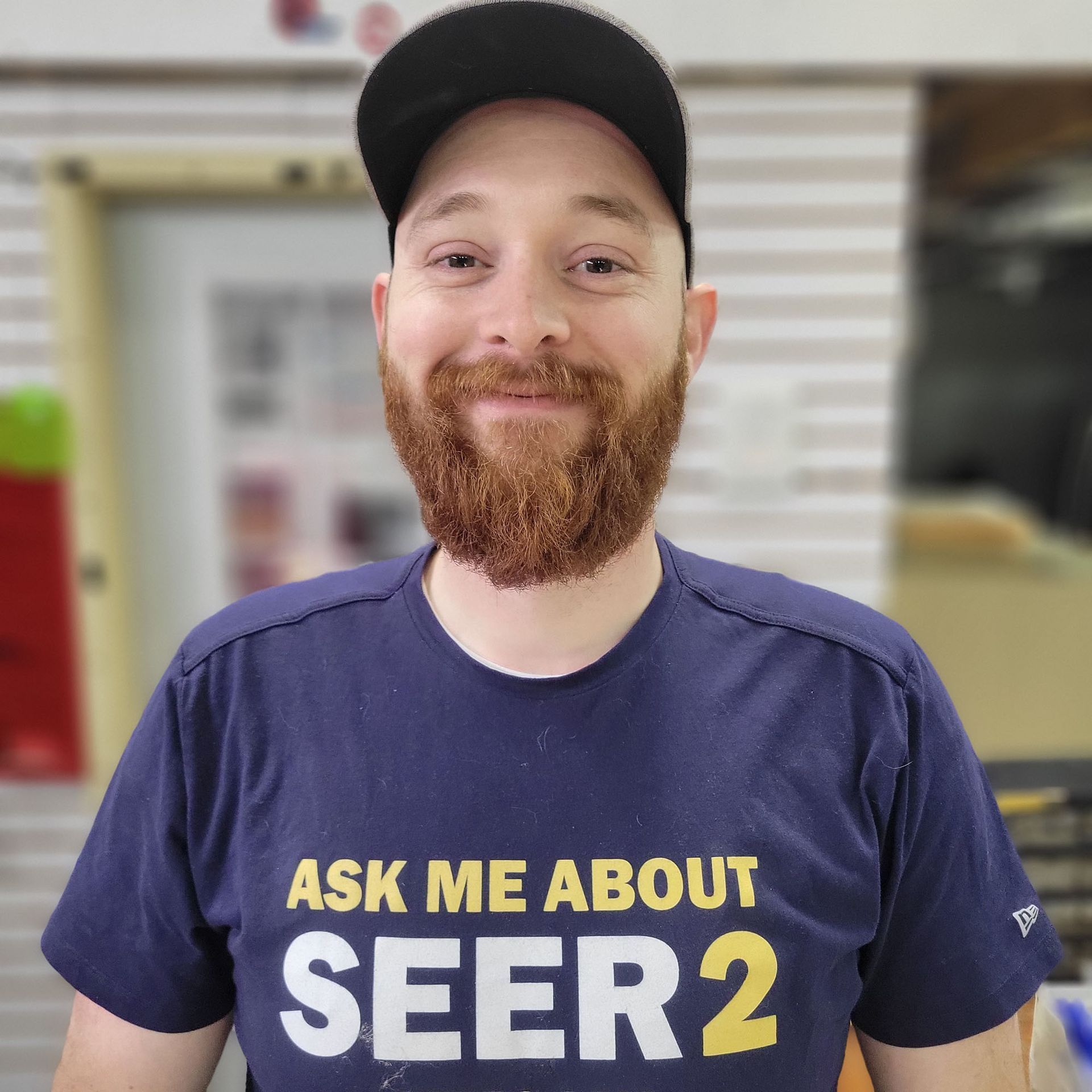 Person smiling in a navy “ASK ME ABOUT SEER2” T-shirt and black cap indoors