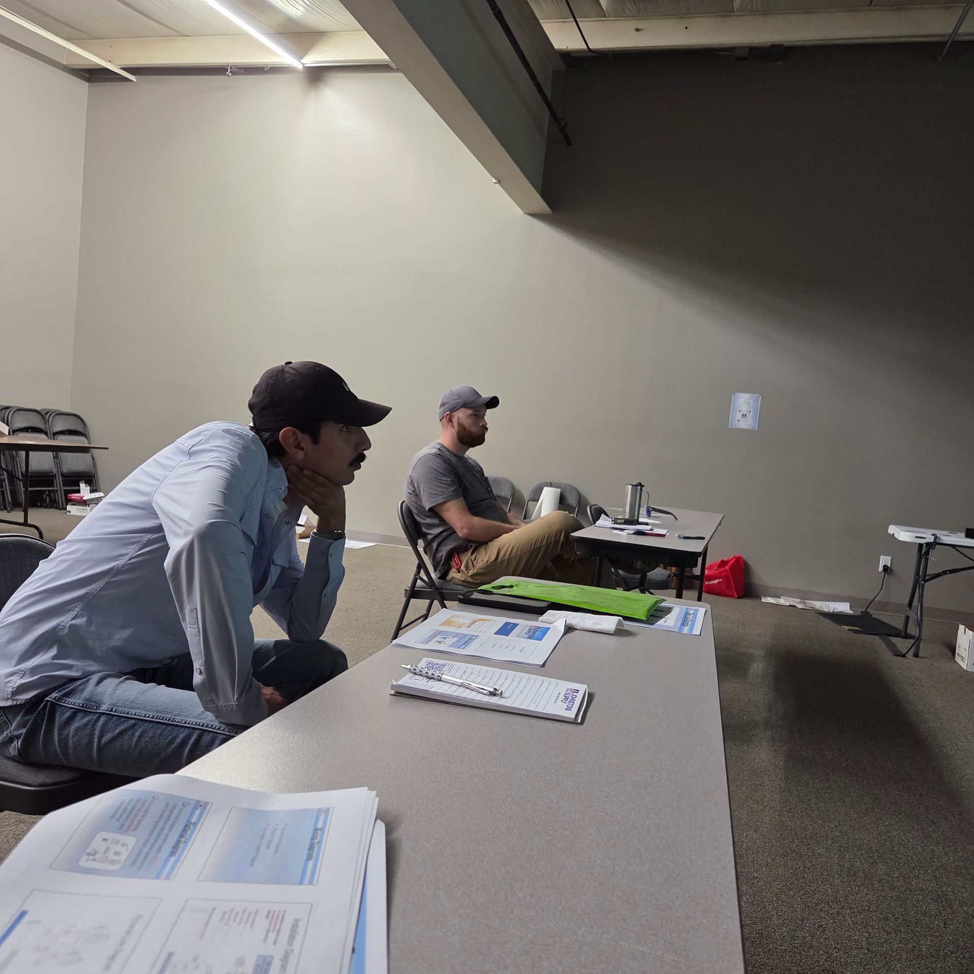 Two people sit at tables in a conference room, looking toward a front wall with papers and laptops nearby.