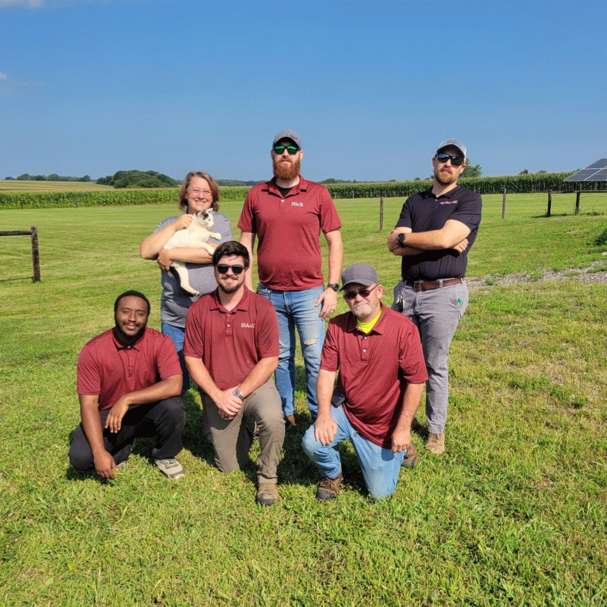 Six people posing together in a sunny grassy field, wearing red shirts and casual clothes.