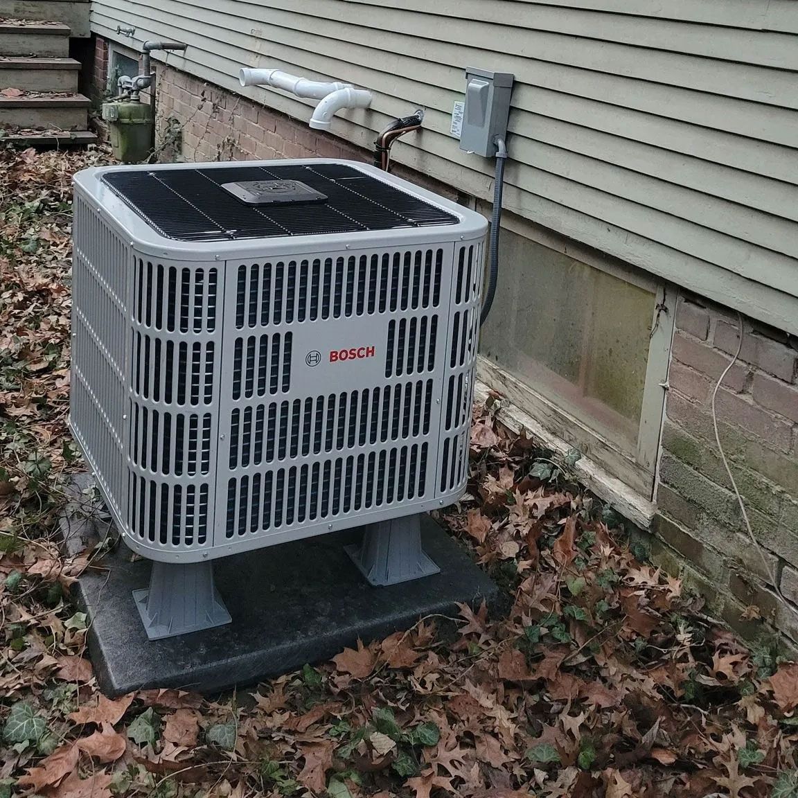 Outdoor Bosch air conditioner unit beside a house wall on a concrete pad, covered with fallen leaves