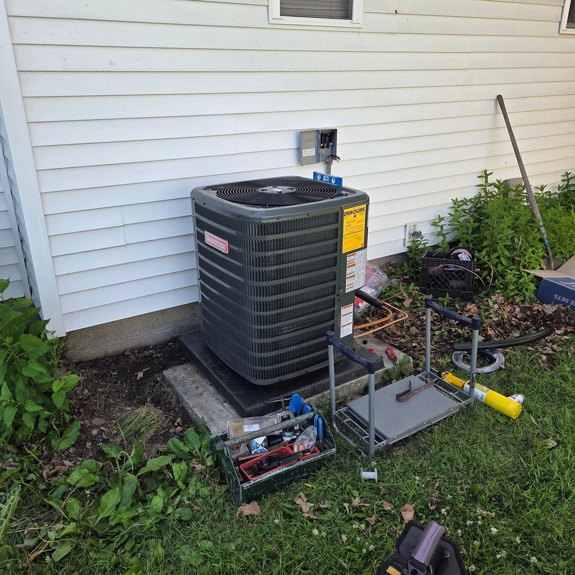 Outdoor HVAC unit beside a white house wall, with exposed piping and tools on a concrete pad.