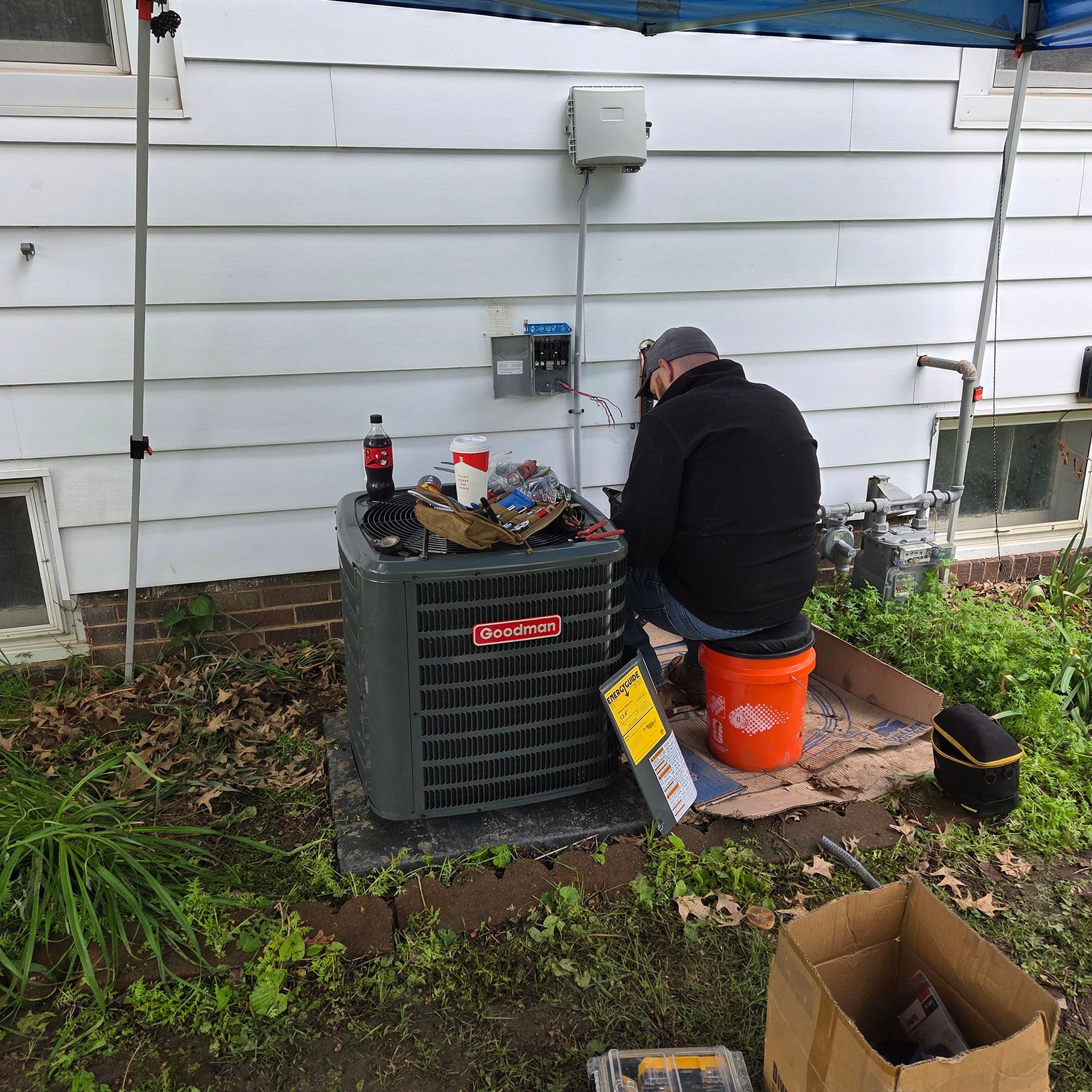 Person kneeling beside an outdoor HVAC unit, working on equipment against a white house wall.