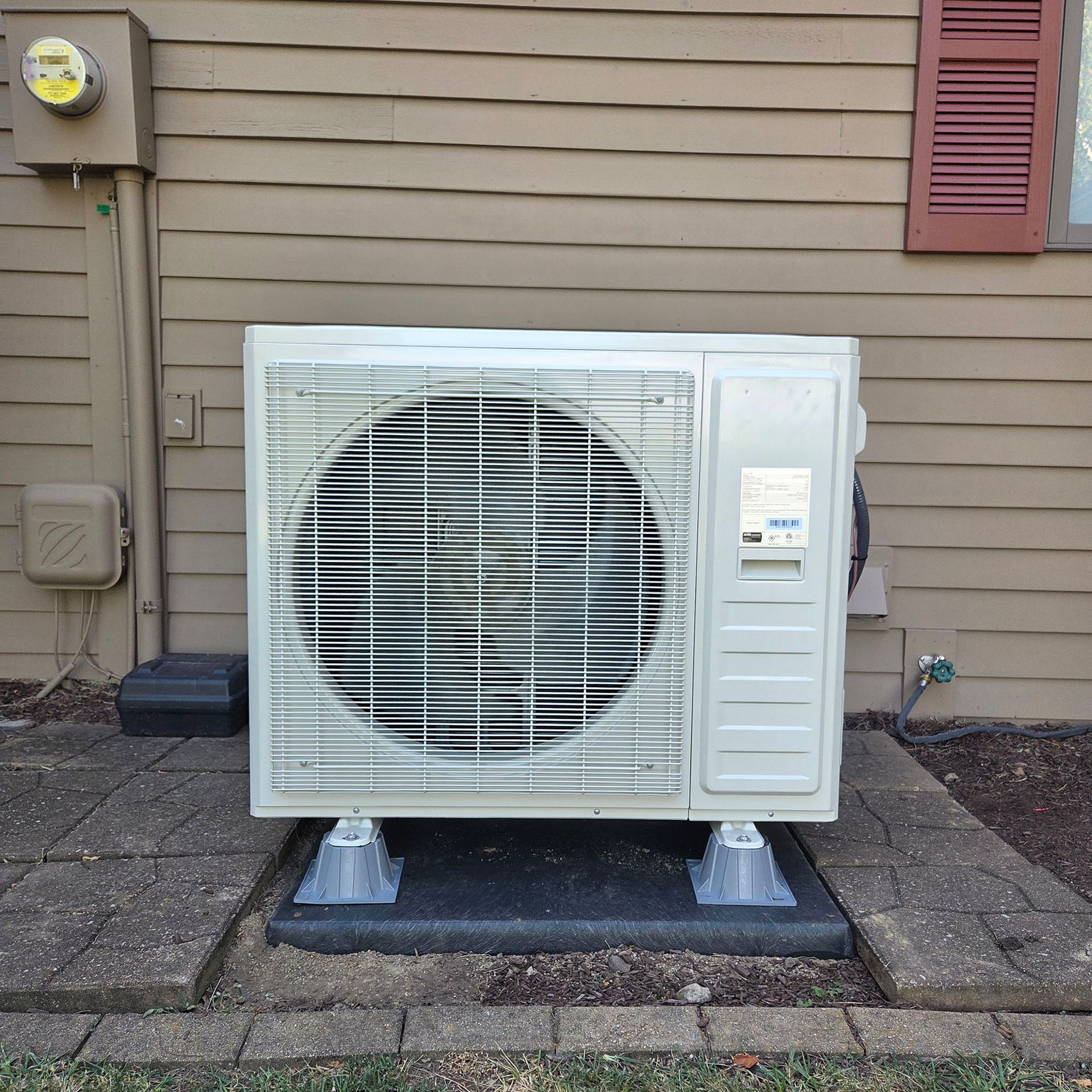 Outdoor air conditioner unit on a concrete pad beside a house with beige siding and a red shutter.