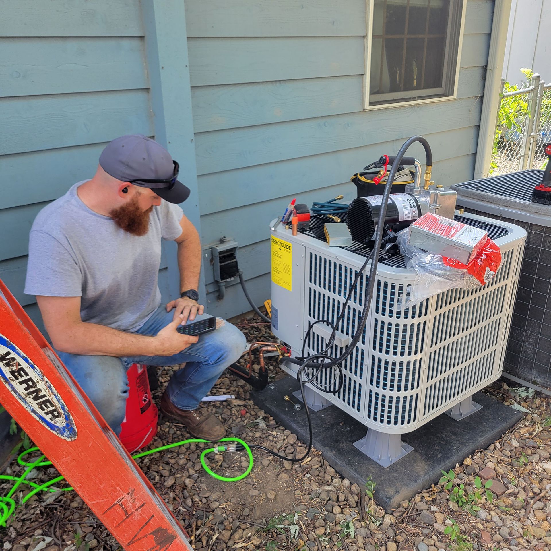 Technician kneeling beside an air compressor unit, checking a hose connection outdoors.
