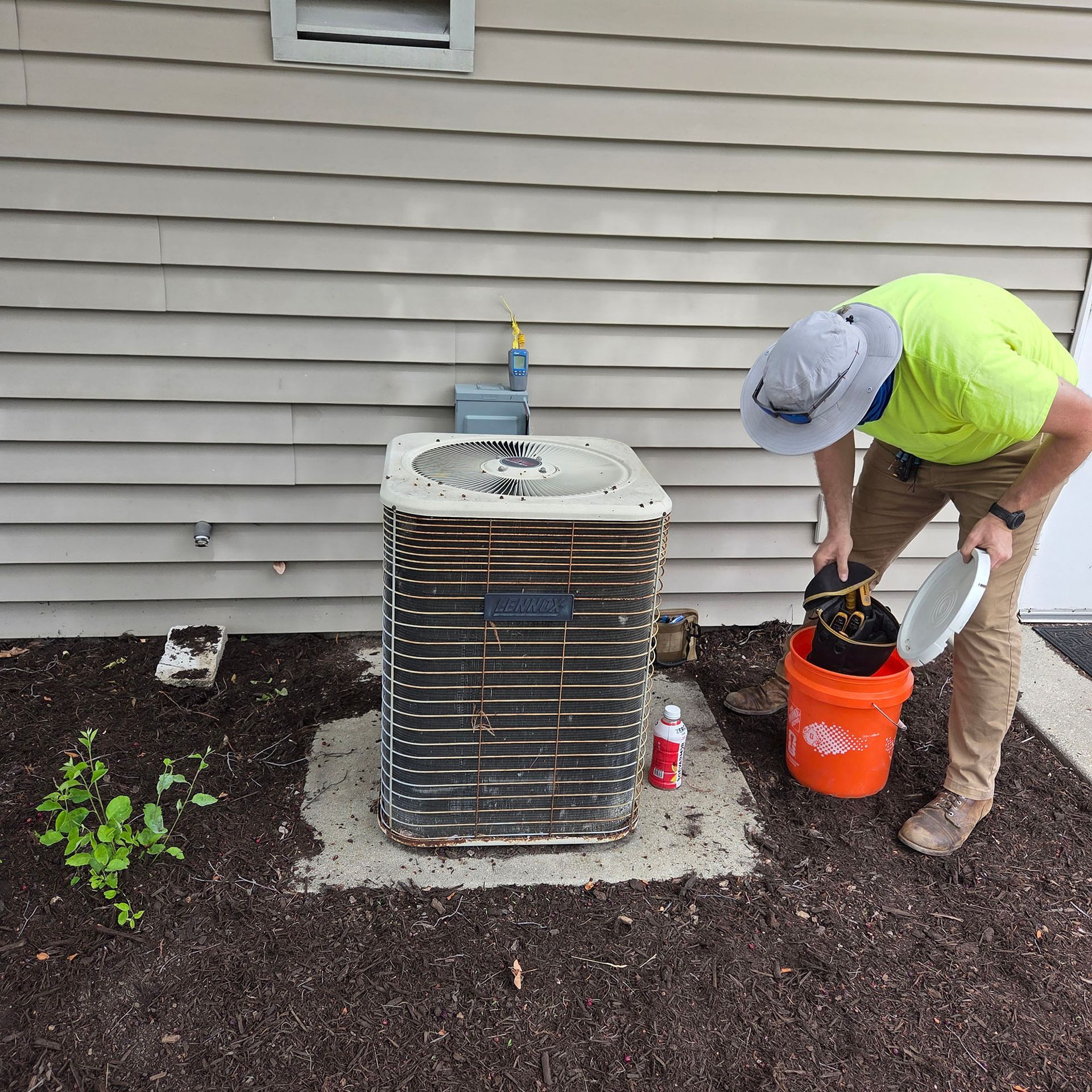 Worker servicing an outdoor HVAC unit beside a house, with tools and an orange bucket.