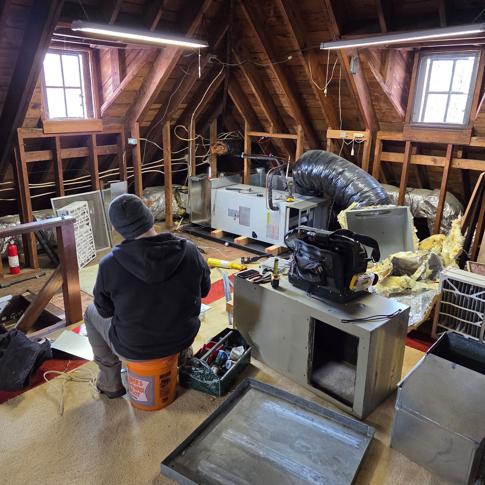 Attic renovation with exposed rafters, ductwork, tools, and a worker seated on a bucket in the foreground