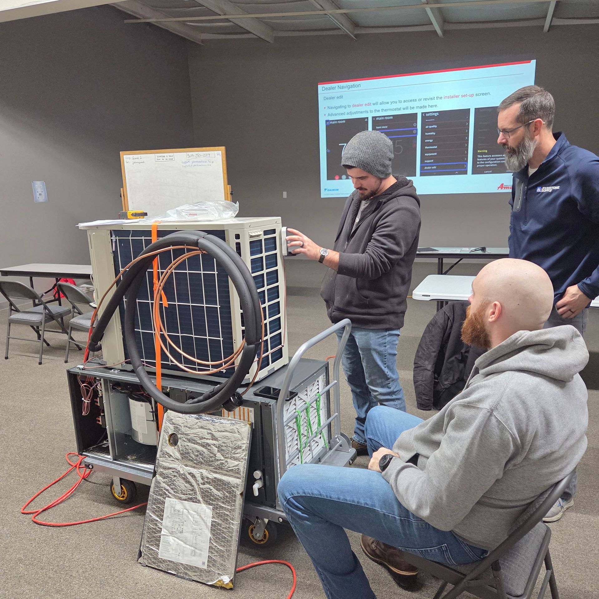 Three people discussing equipment beside a wheeled apparatus in a workshop, with a presentation projected behind them.