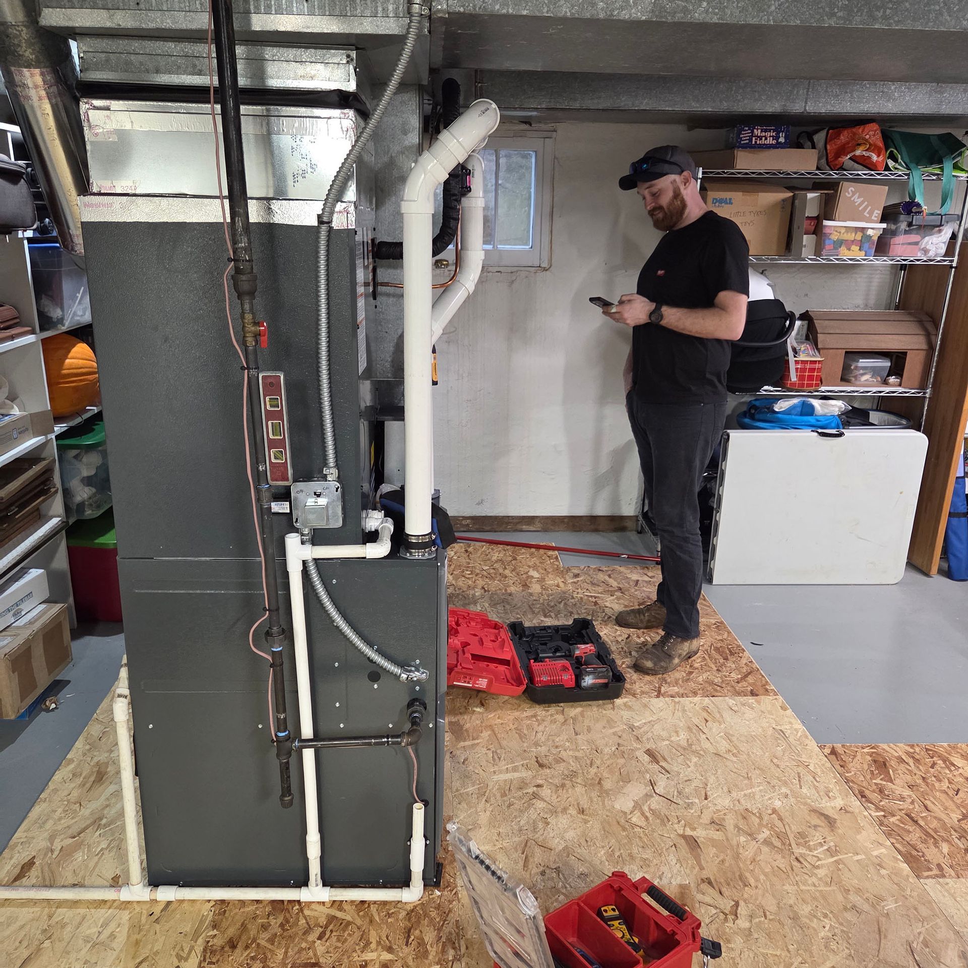 Man standing on plywood floor in a utility room, looking at a phone beside exposed HVAC equipment and tools.