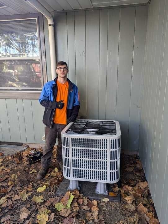 Person standing beside an outdoor HVAC unit under a porch, wearing a blue jacket and orange shirt