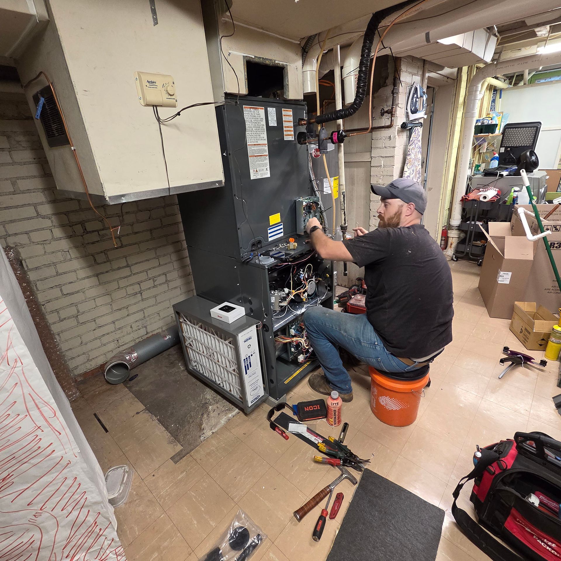 Technician kneeling beside HVAC unit, working with tools in a utility room cluttered with boxes and equipment.