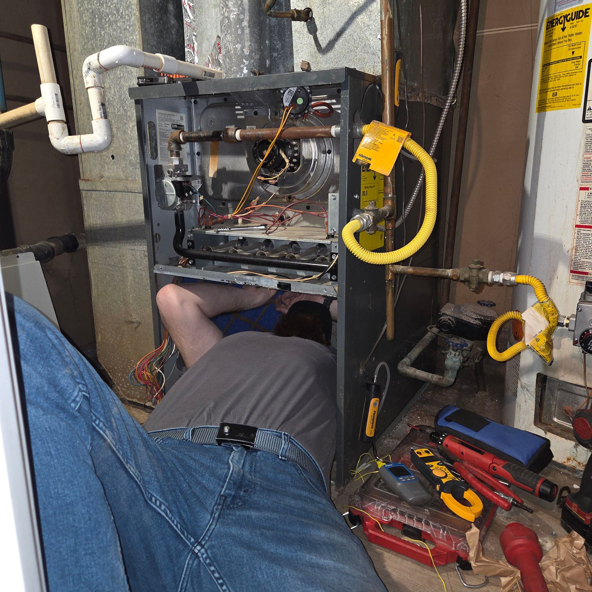 Technician working inside an open furnace cabinet in a basement, with tools scattered nearby.