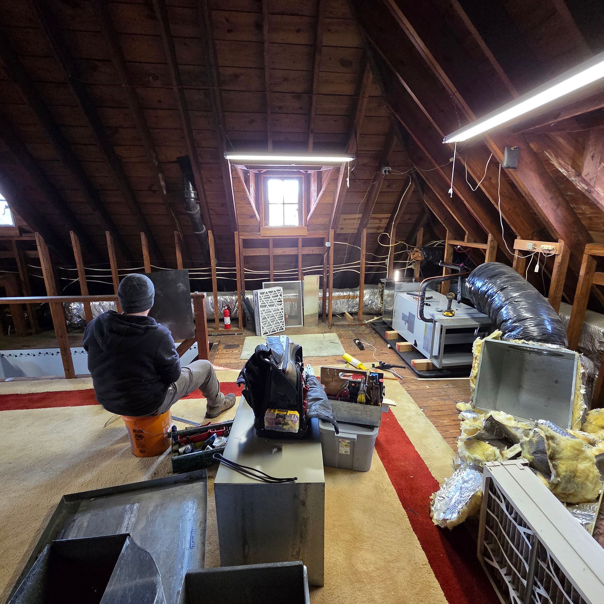 Person working in an attic with exposed beams, insulation, boxes, and HVAC equipment scattered around.