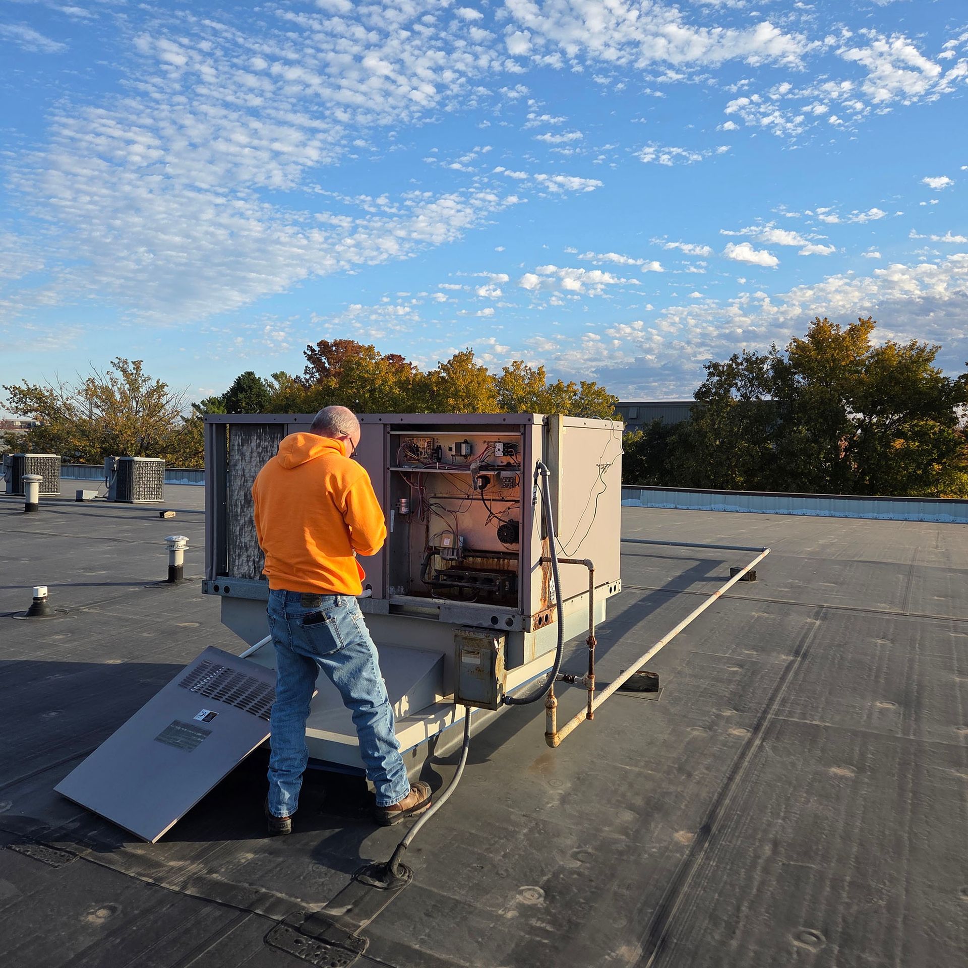 Man in orange shirt inspecting rooftop HVAC unit under a blue sky