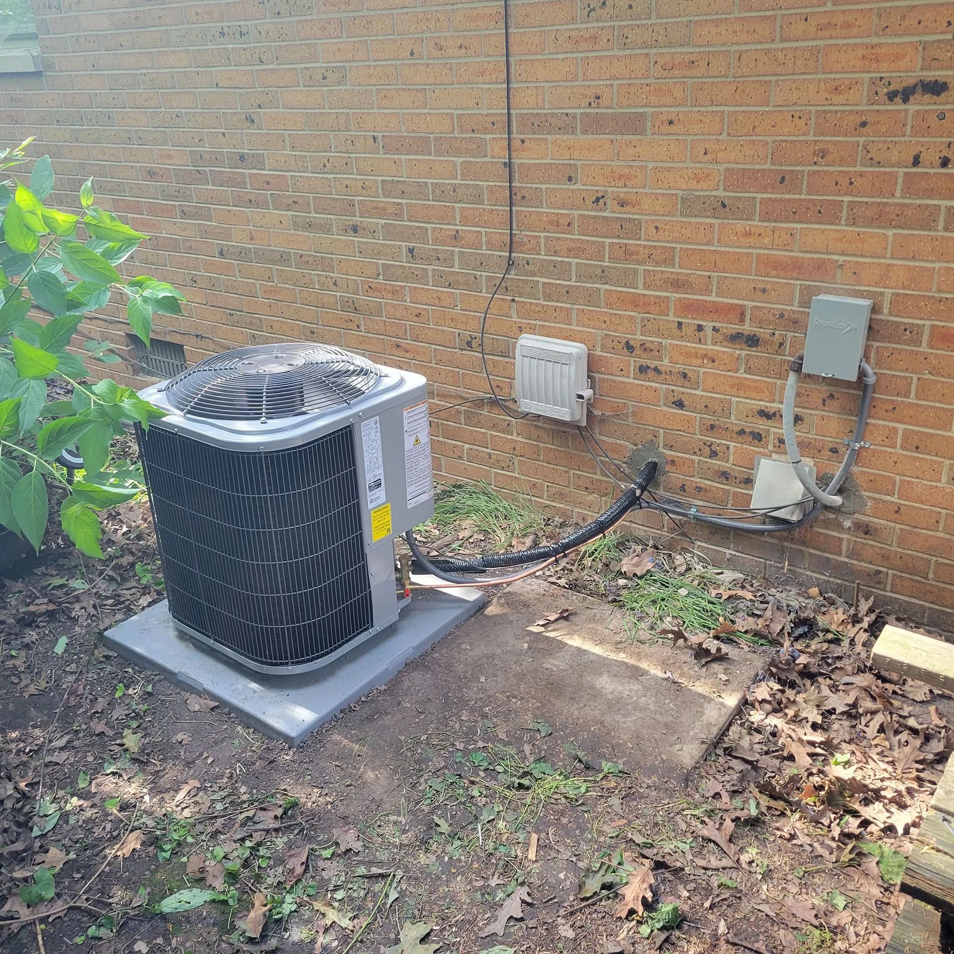 Outdoor air conditioner unit beside a brick wall with electrical boxes and fallen leaves on the ground