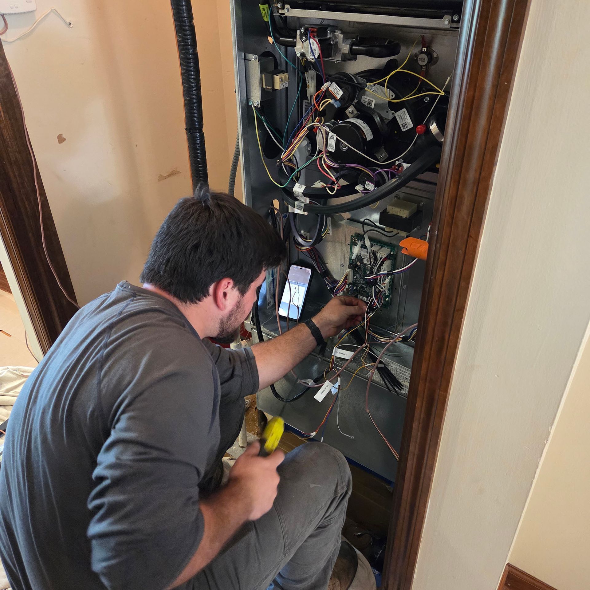 Technician working inside an open wall cabinet, using a smartphone and yellow tool amid tangled wiring.