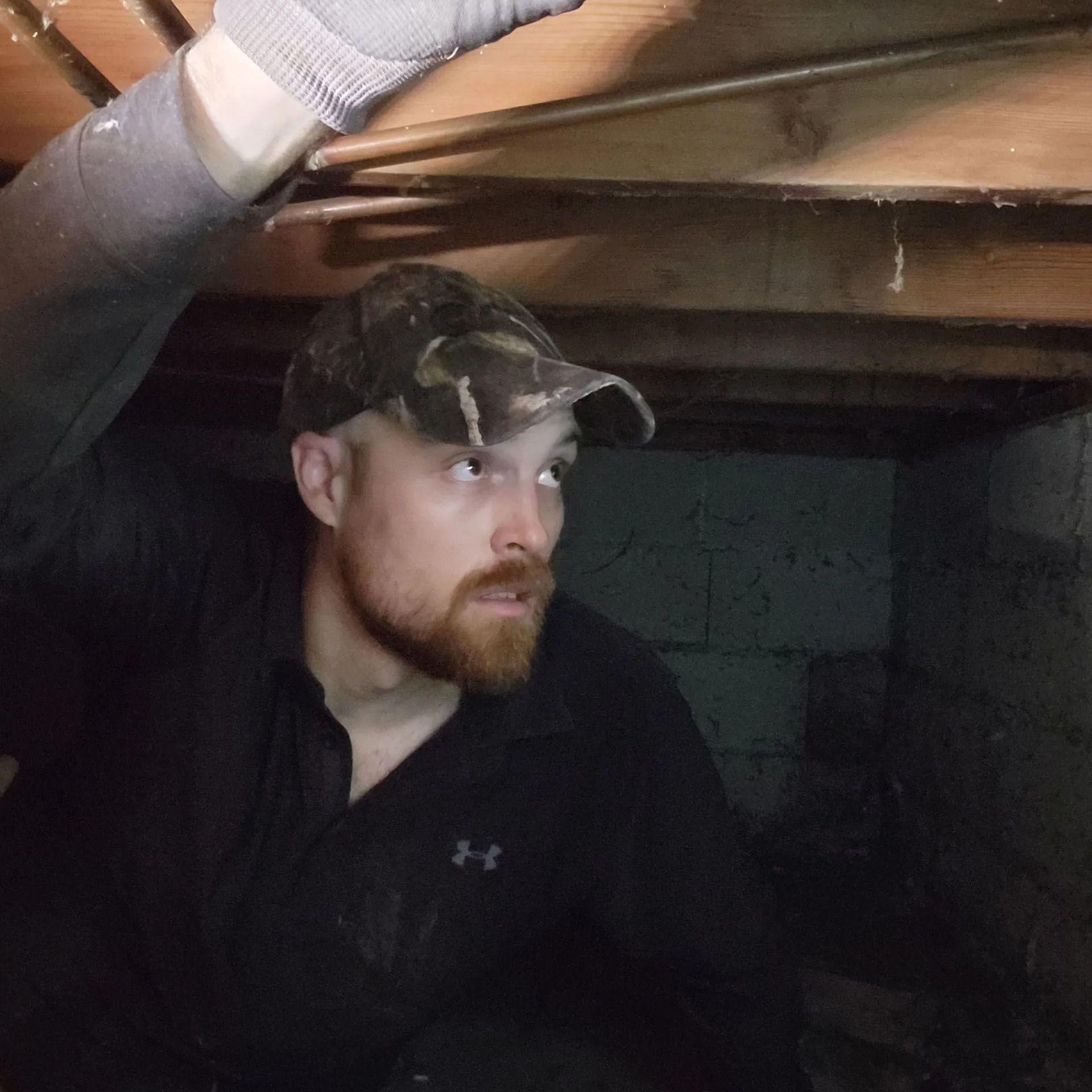 Person under a low wooden beam in a dark crawlspace, looking to the side, wearing a cap and dark shirt.