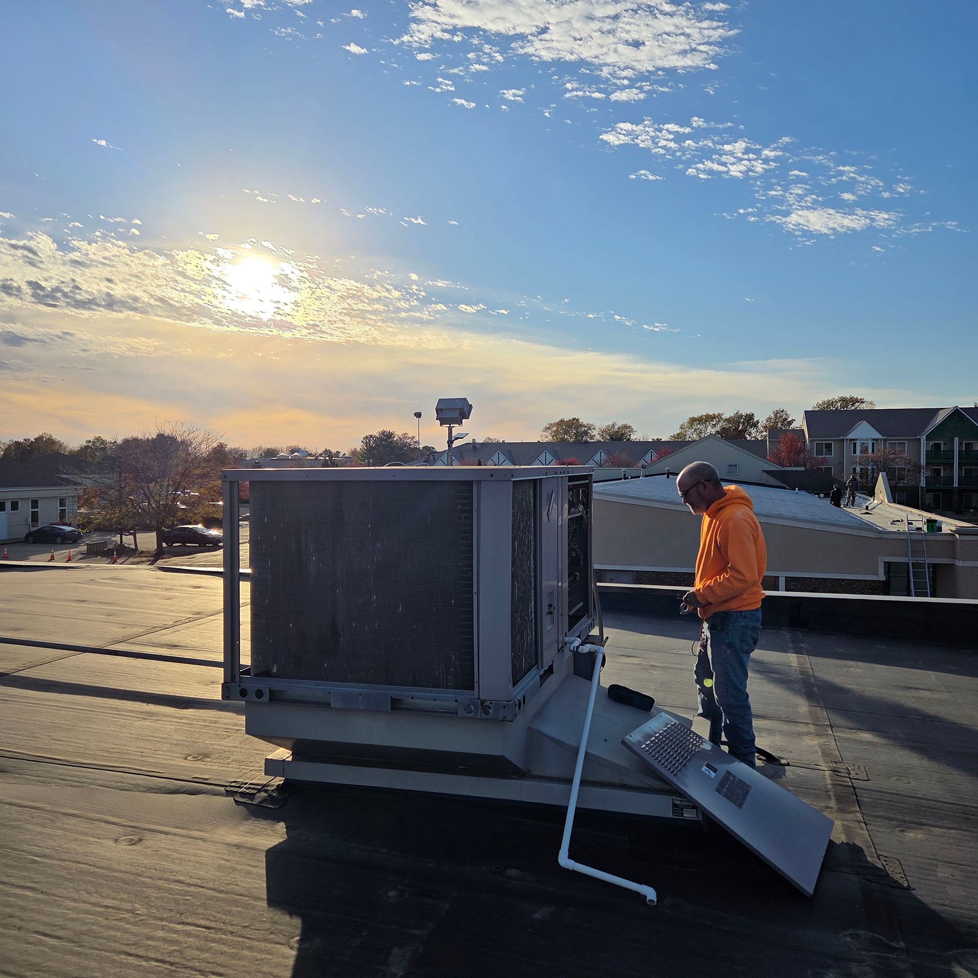 Worker in orange jacket on a rooftop beside a large HVAC unit at sunset.