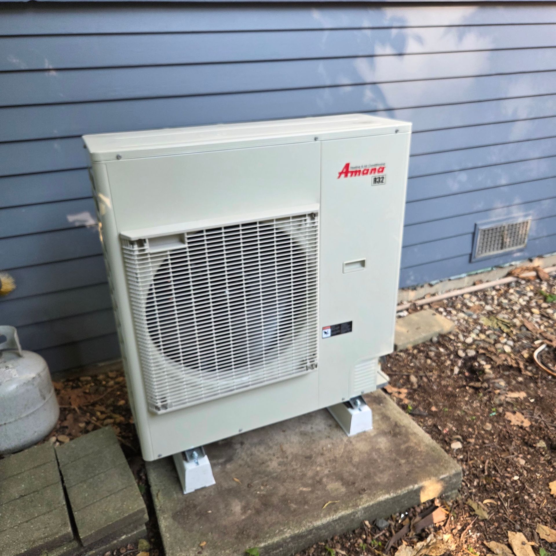 Outdoor Amana HVAC condenser unit beside a house with blue siding on a concrete pad