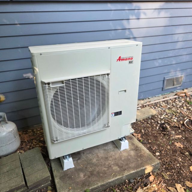 Outdoor Amana HVAC condenser unit beside a house with blue siding on a concrete pad