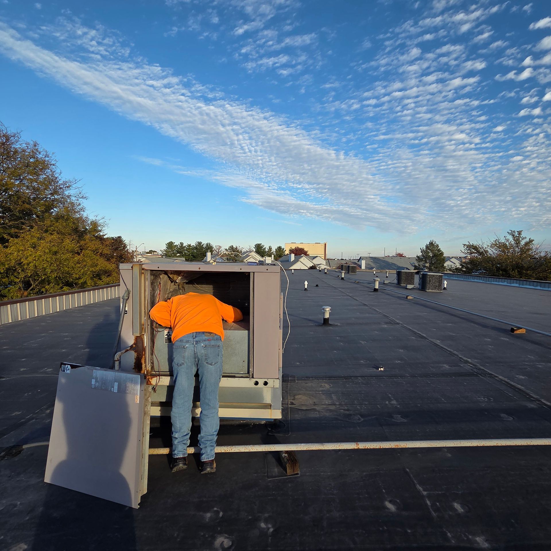Worker in orange shirt inspecting rooftop HVAC unit under a blue sky