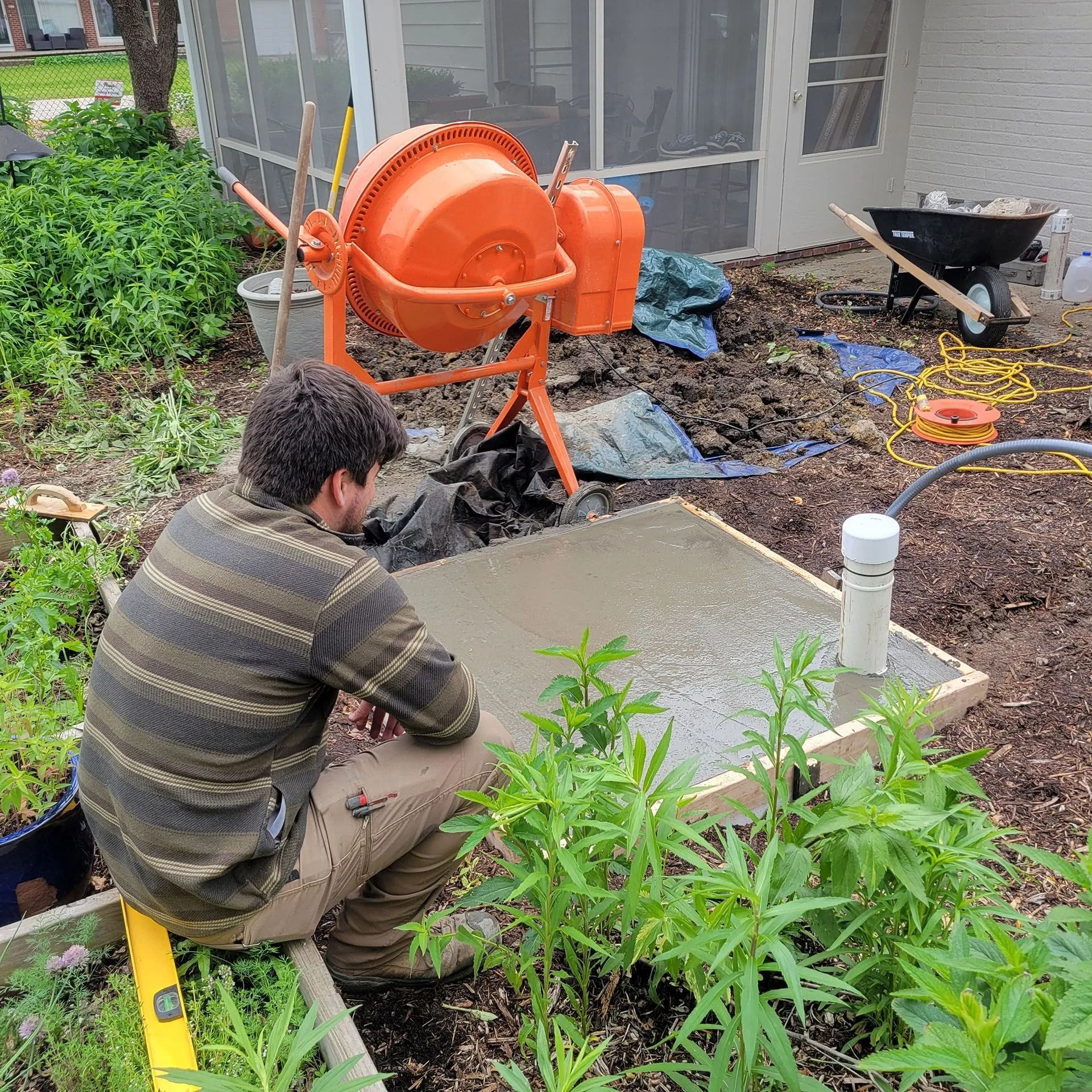 Worker pouring concrete beside an orange cement mixer in a garden construction site.