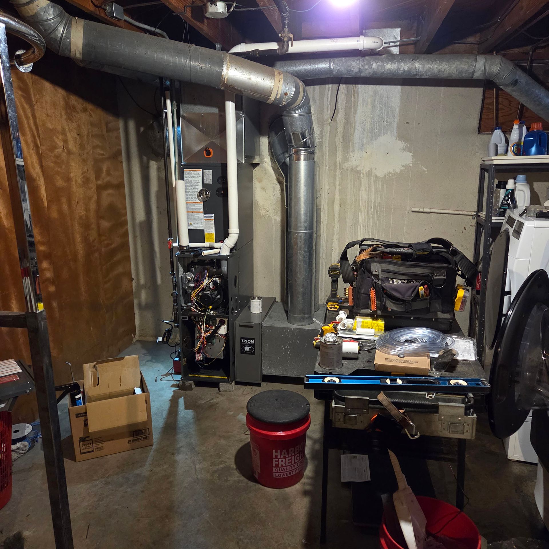 Basement workshop with exposed ducts, workbench, tools, and a red bucket on the floor.