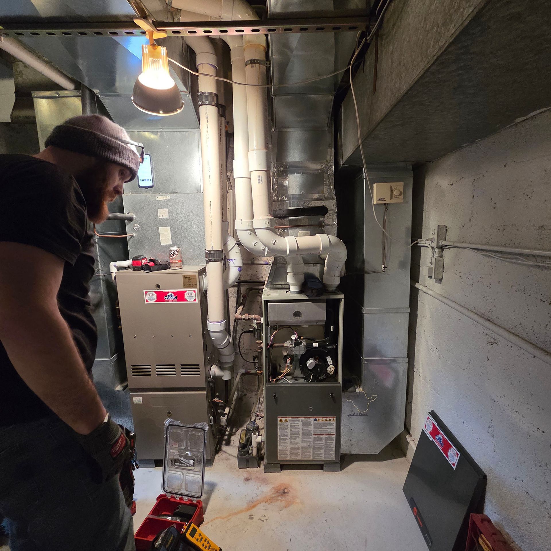 Man inspecting furnace and ductwork in a cramped basement utility room with exposed pipes and hanging light.