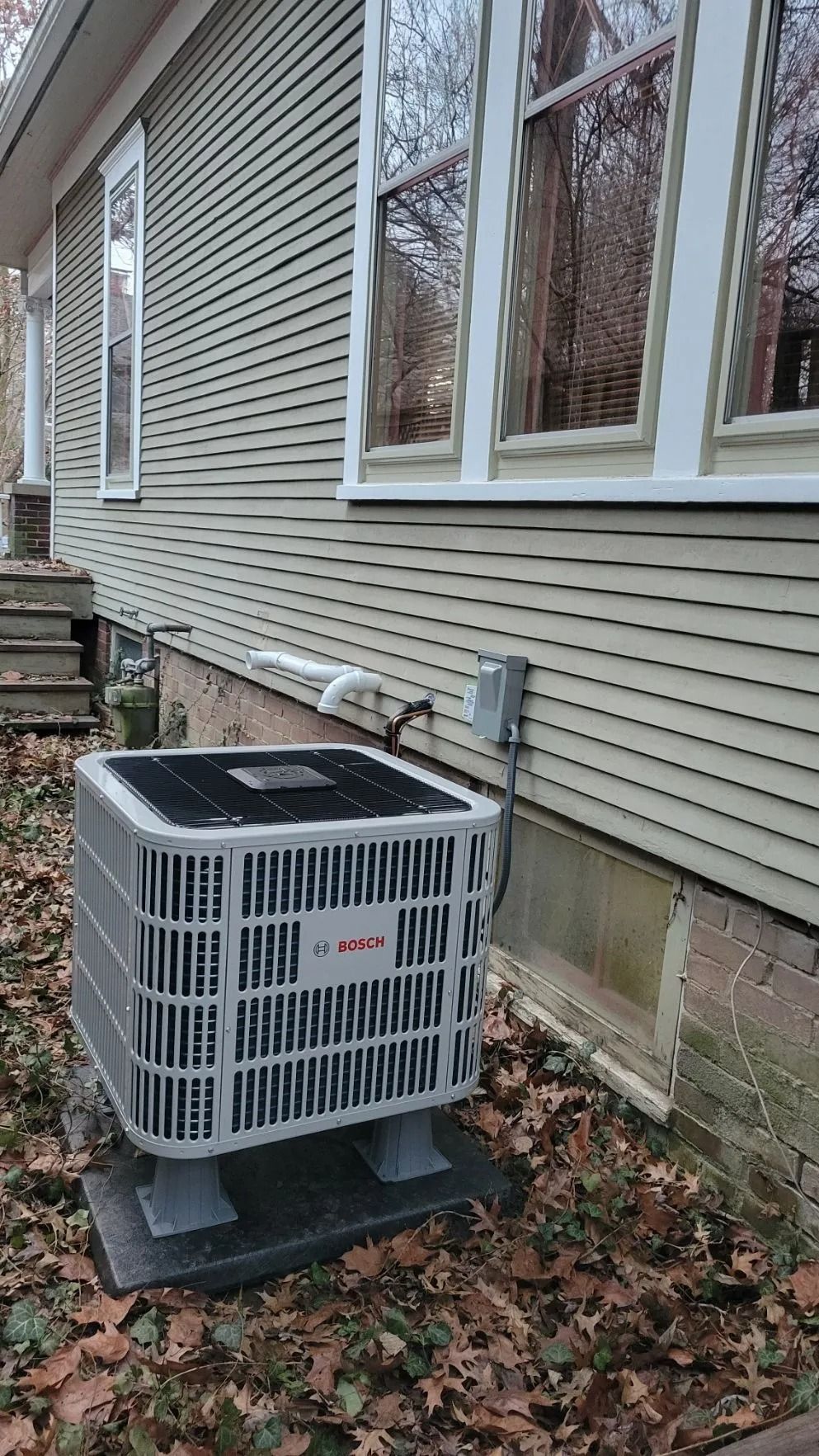 Outdoor HVAC unit beside a beige house with vinyl siding and windows, surrounded by fallen leaves