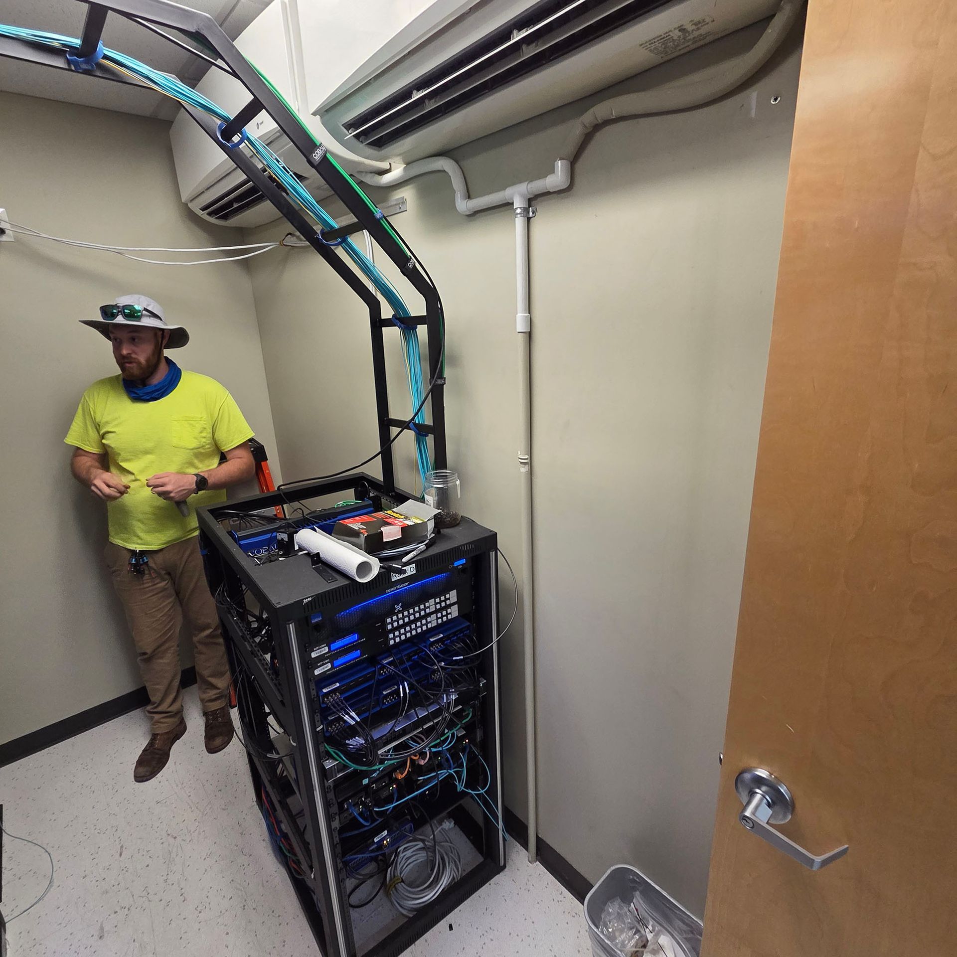 Technician standing beside a server rack in a utility room with overhead cable trays and an open door.