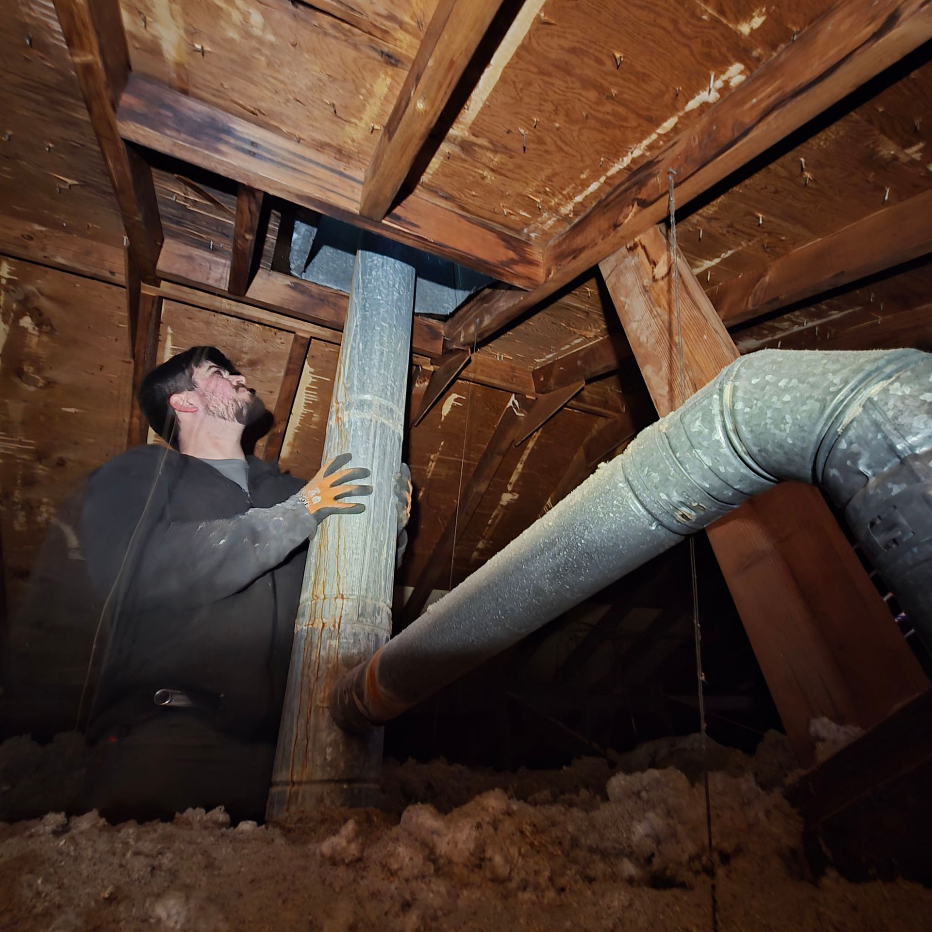 Worker inspecting exposed ductwork and pipes in a crawlspace beneath wooden floor joists