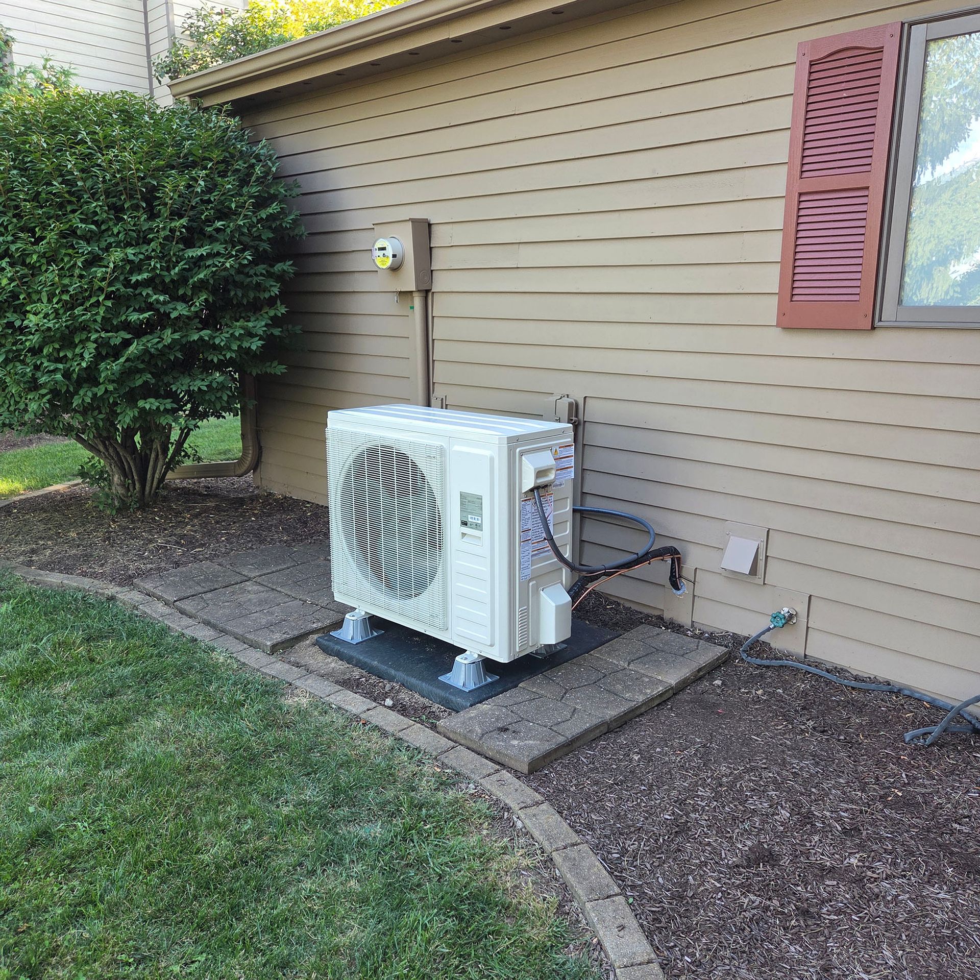 Outdoor HVAC heat pump unit beside a tan house wall, near a window and lawn.