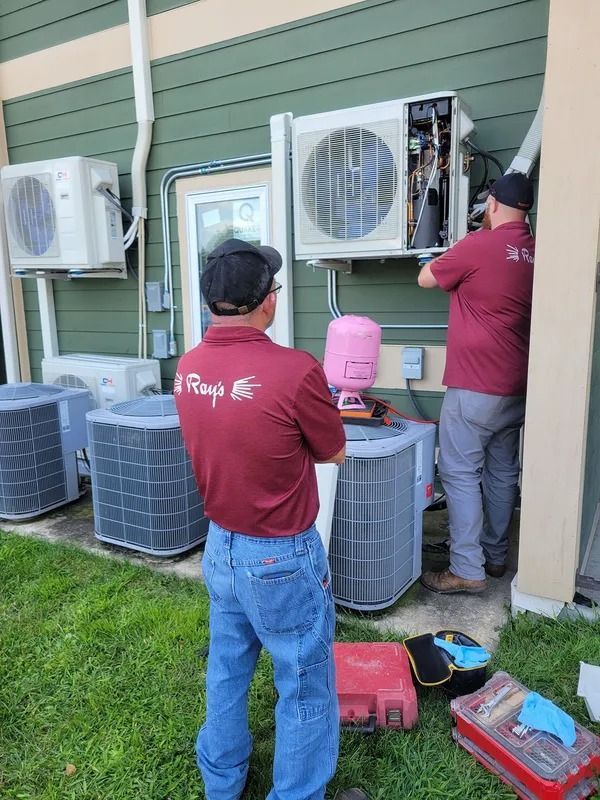 Two technicians service outdoor air-conditioning units against a green house wall.