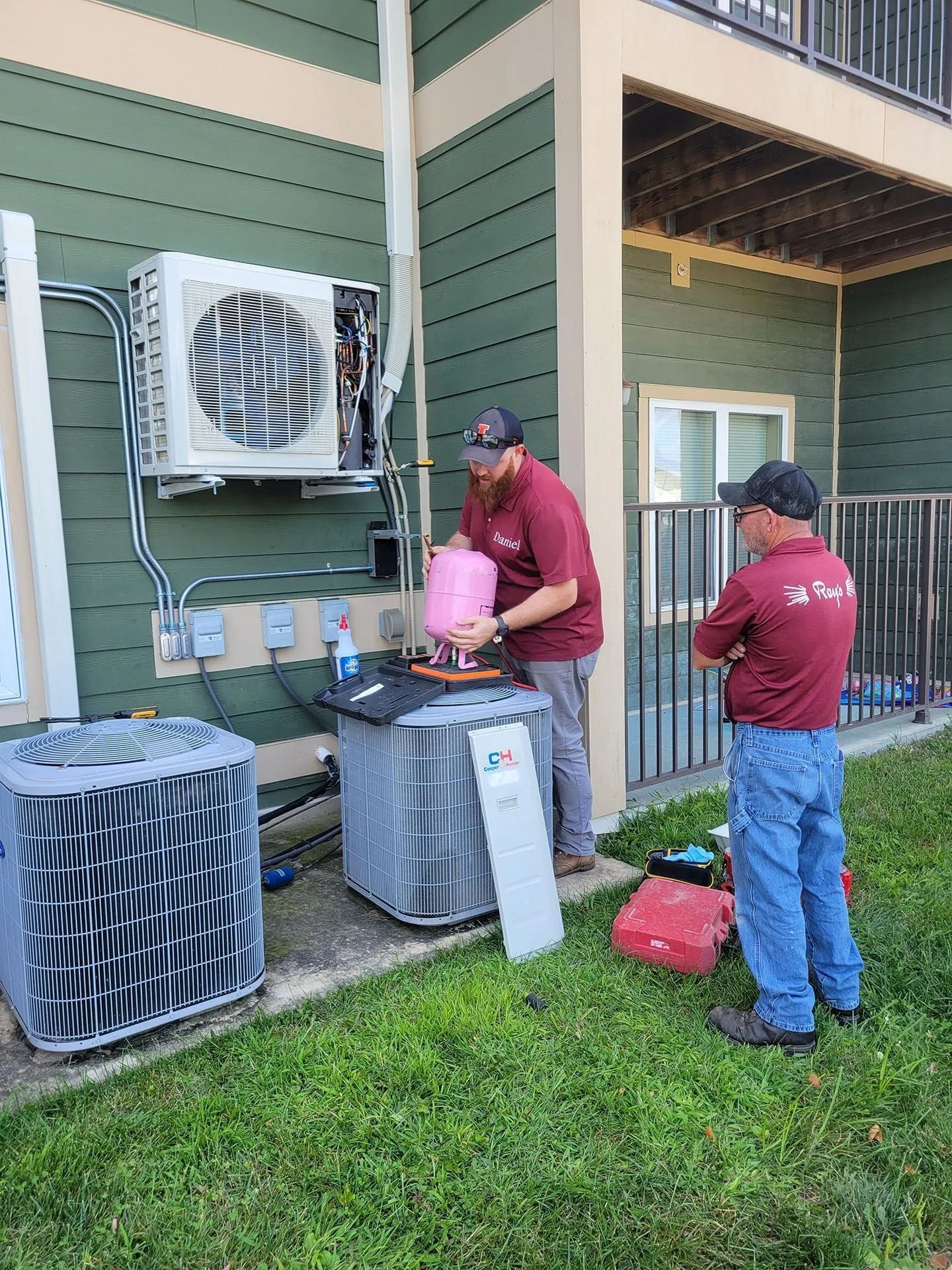 Two workers inspect outdoor HVAC units beside a green apartment building.