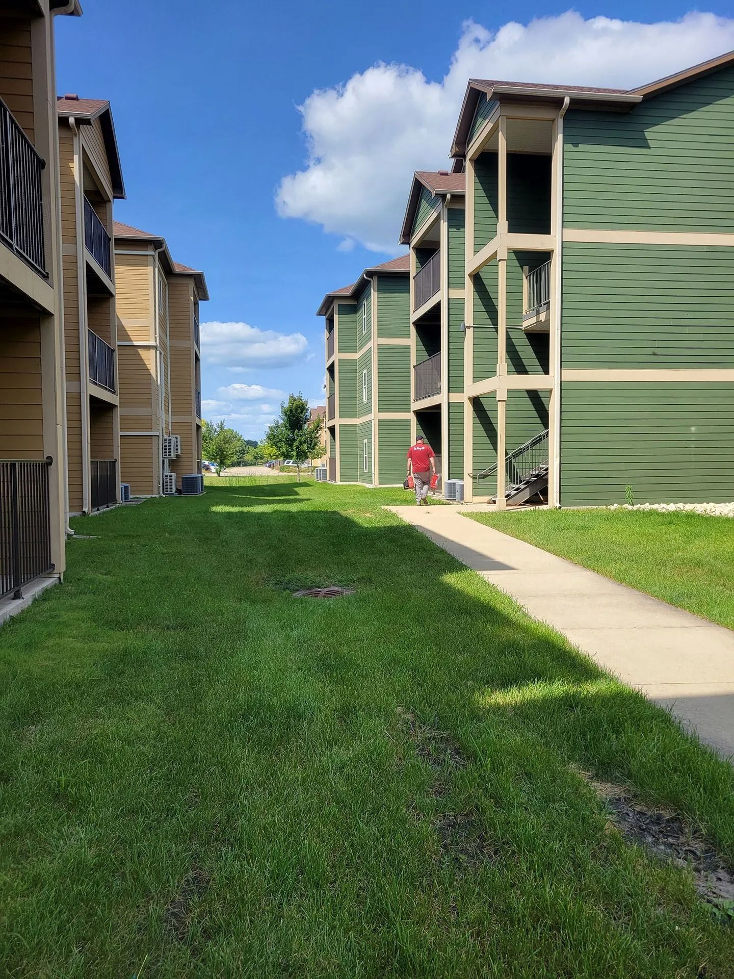 Apartment buildings with green siding beside a grassy courtyard and sidewalk under a blue sky