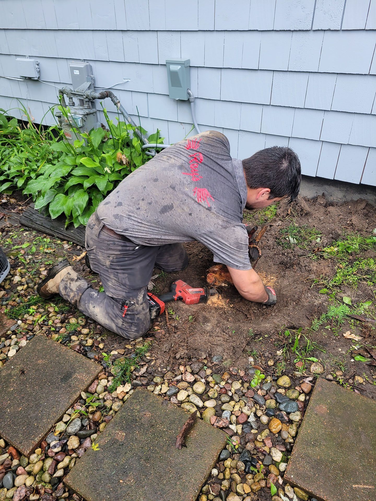 Person kneeling in a garden bed, digging in the soil beside a house wall and pavers.
