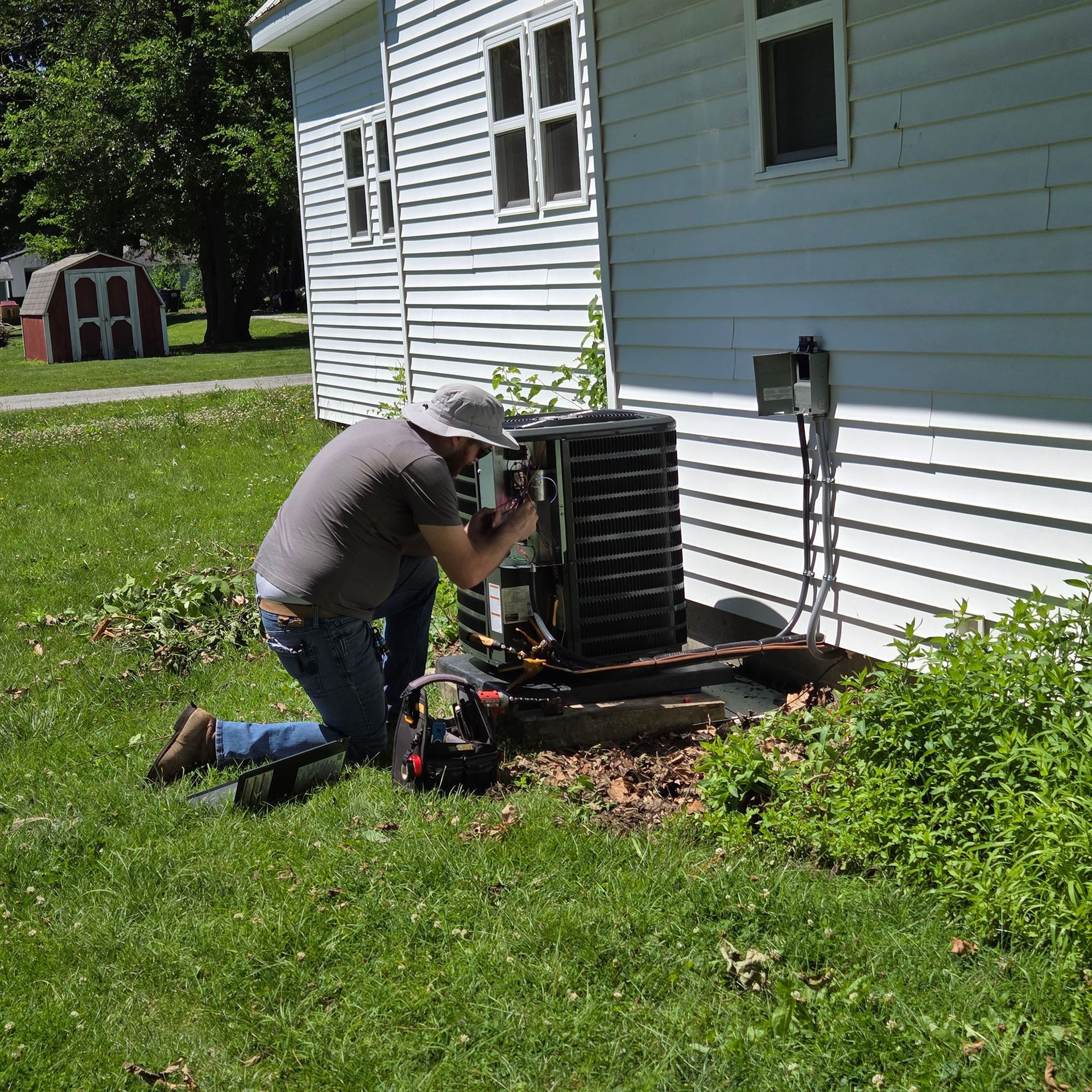 Technician kneeling beside a house, inspecting an outdoor HVAC unit in the grass.
