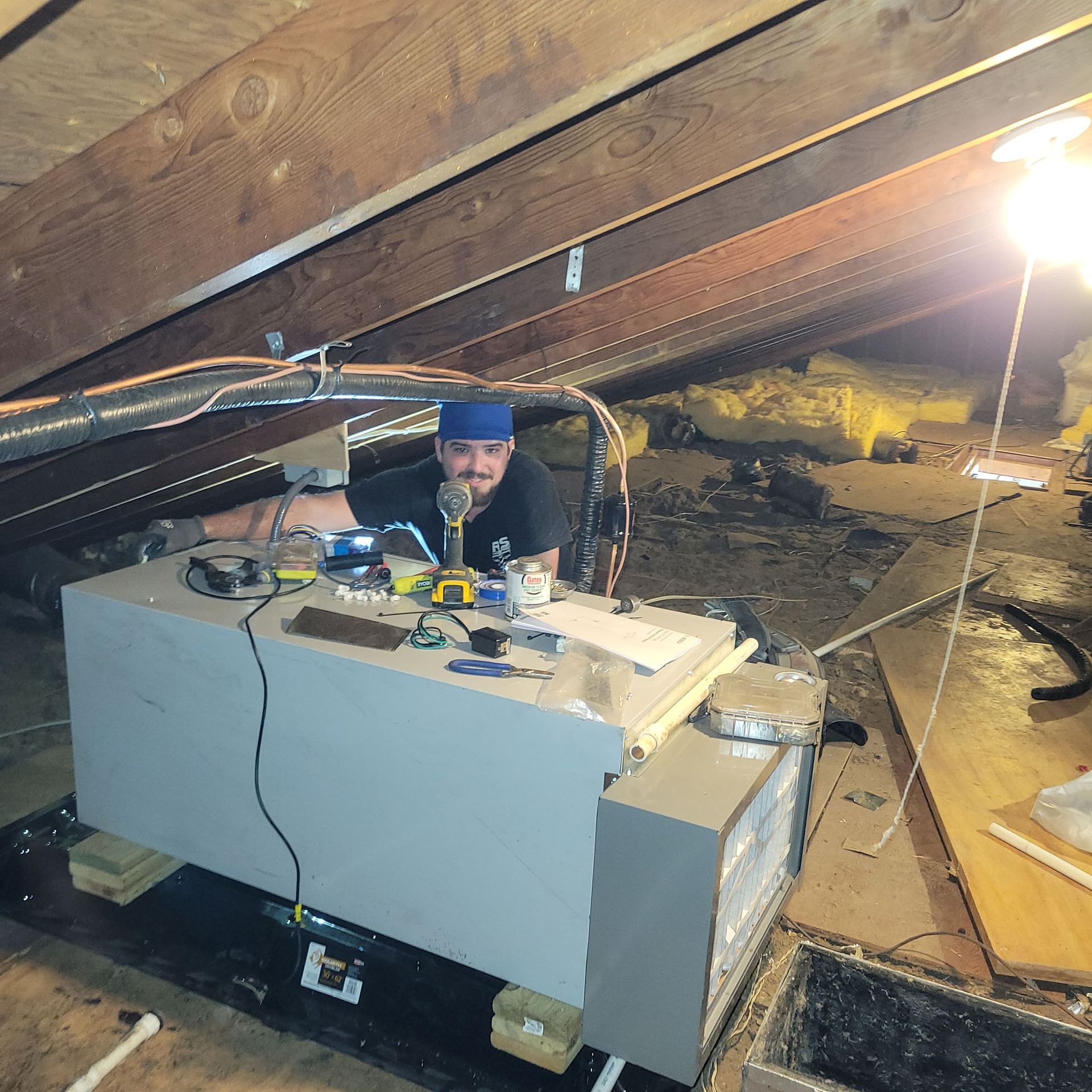 Person crouched under a slanted roof, working on a large gray machine in a dim attic space.