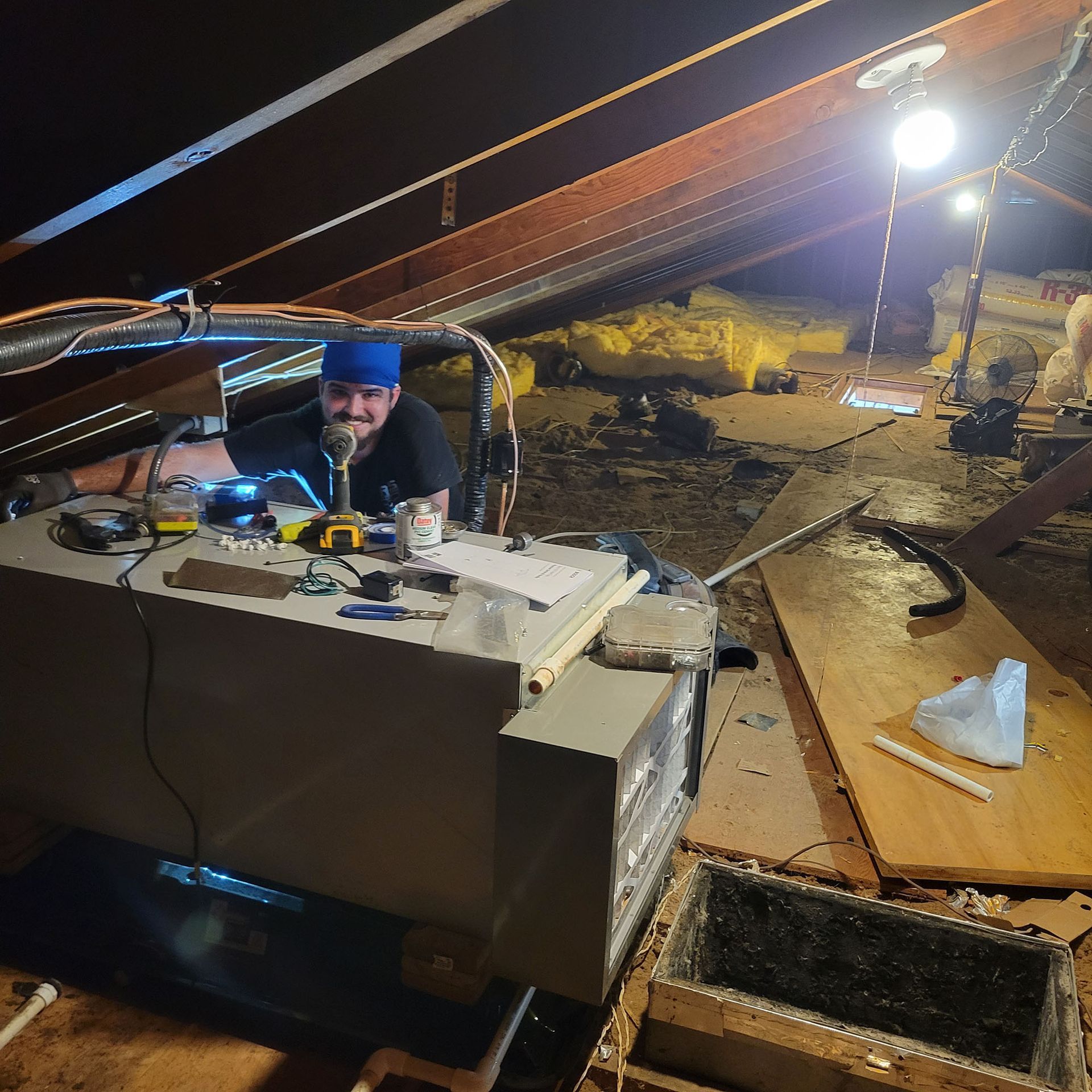 Person working on equipment inside a dim attic with exposed beams and a bright work light.