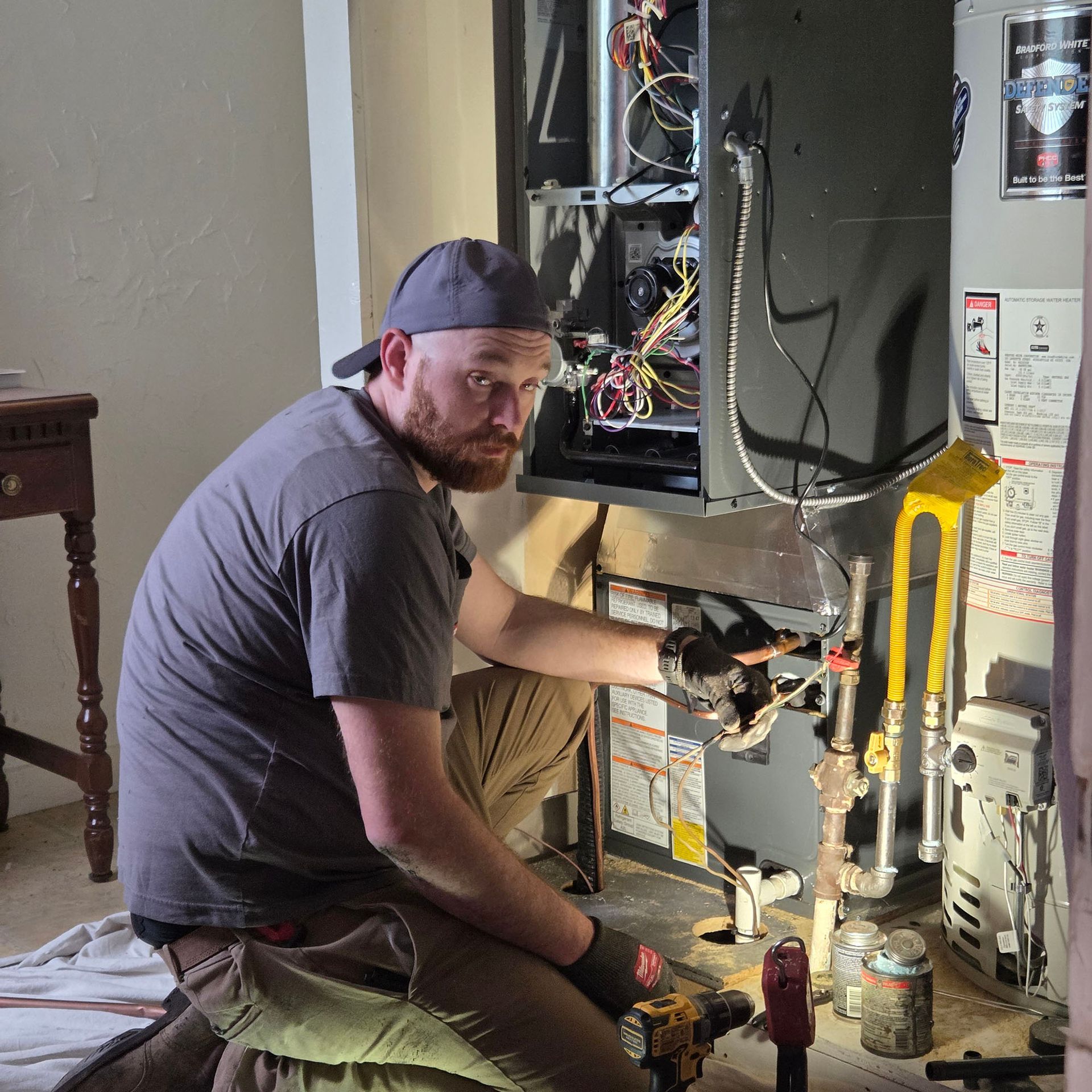 Person kneeling beside an open electrical panel, using tools in a utility room with exposed wiring and pipes