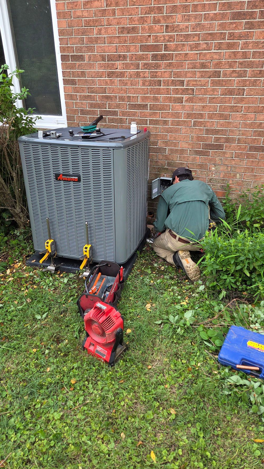 Technician kneels beside outdoor HVAC unit, working on equipment by a brick wall with tools on the grass.