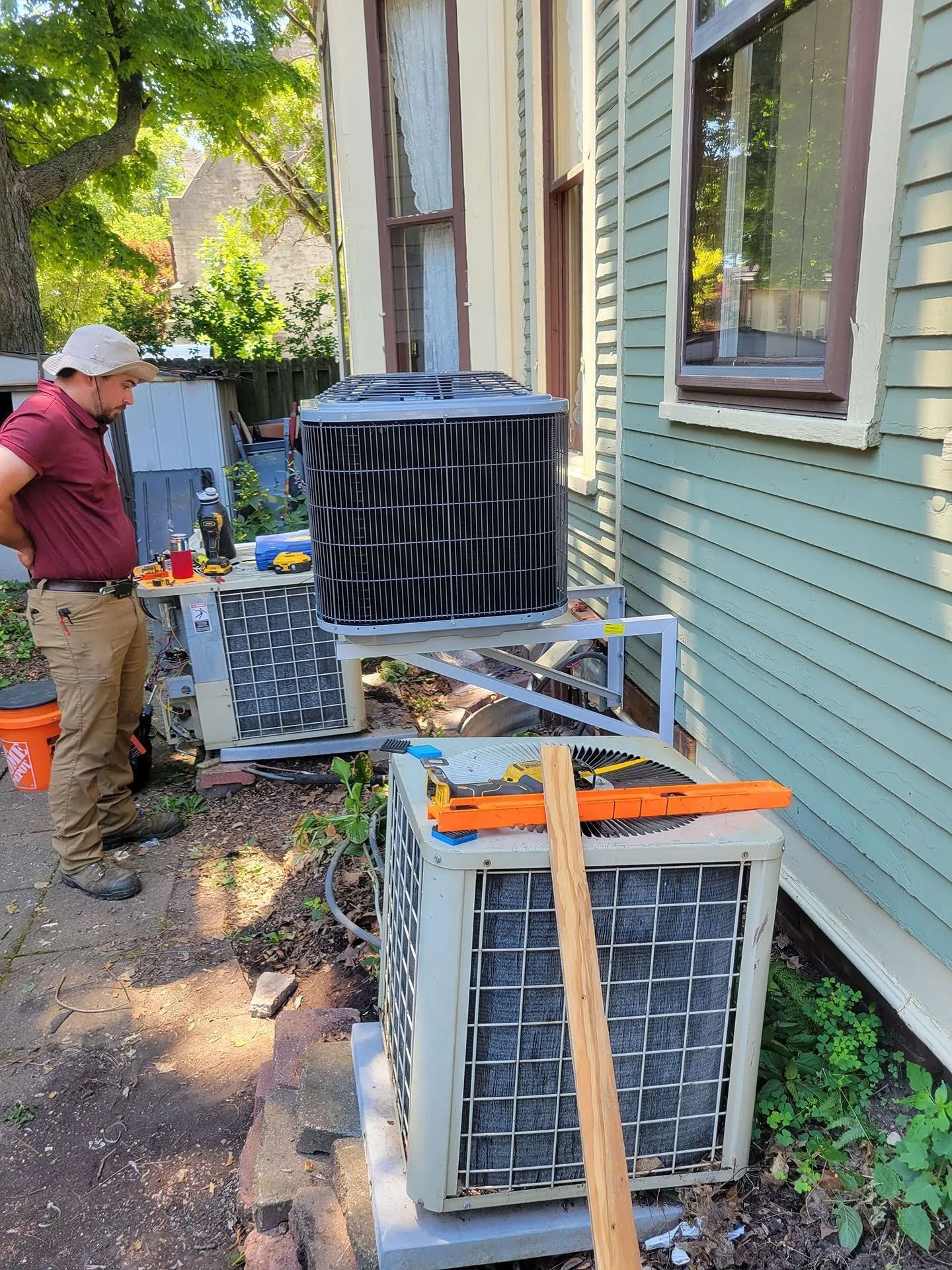 Man working beside two outdoor HVAC units near a house, with tools and equipment on a side yard path