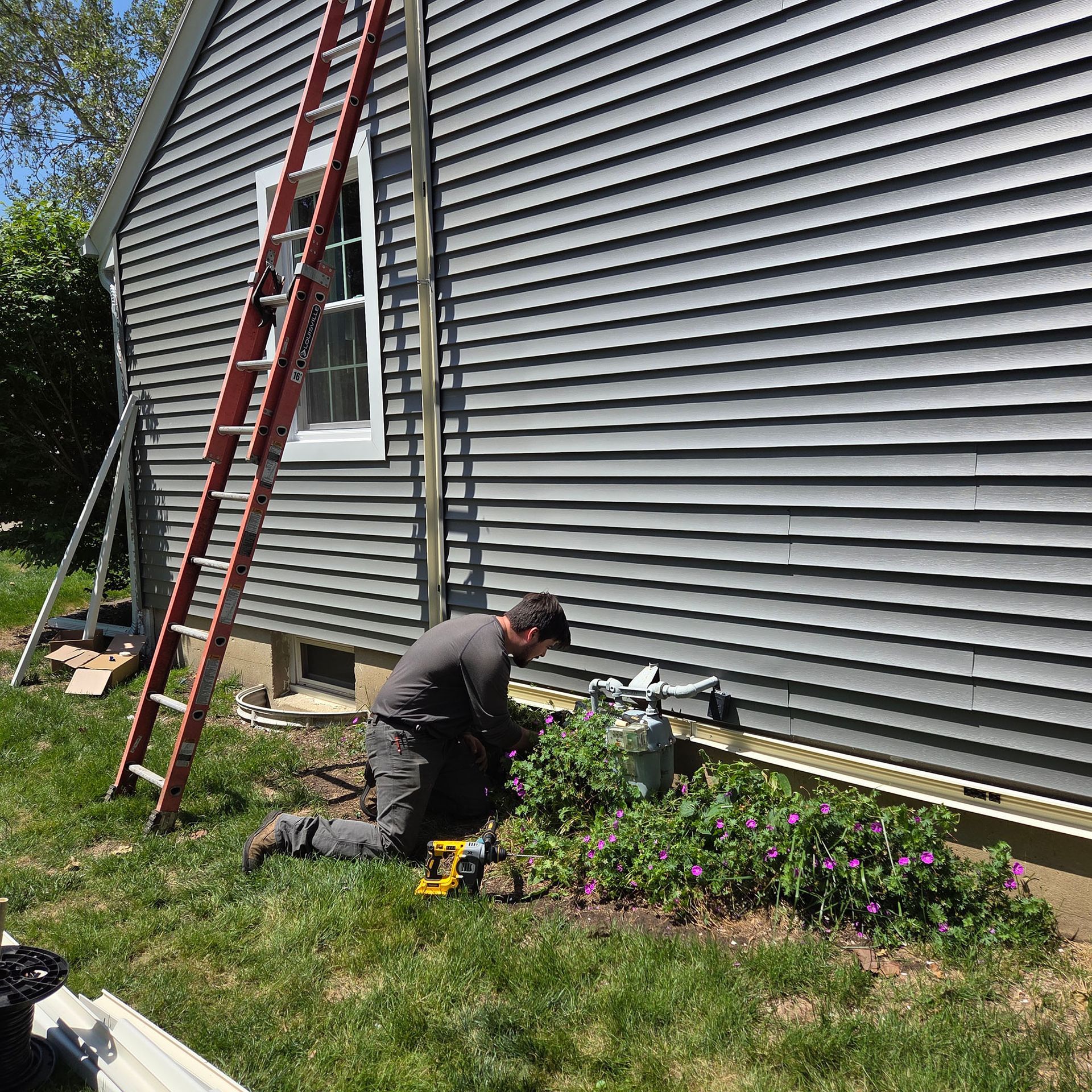 Worker kneels by a house wall, using tools beside a red ladder and garden bed.