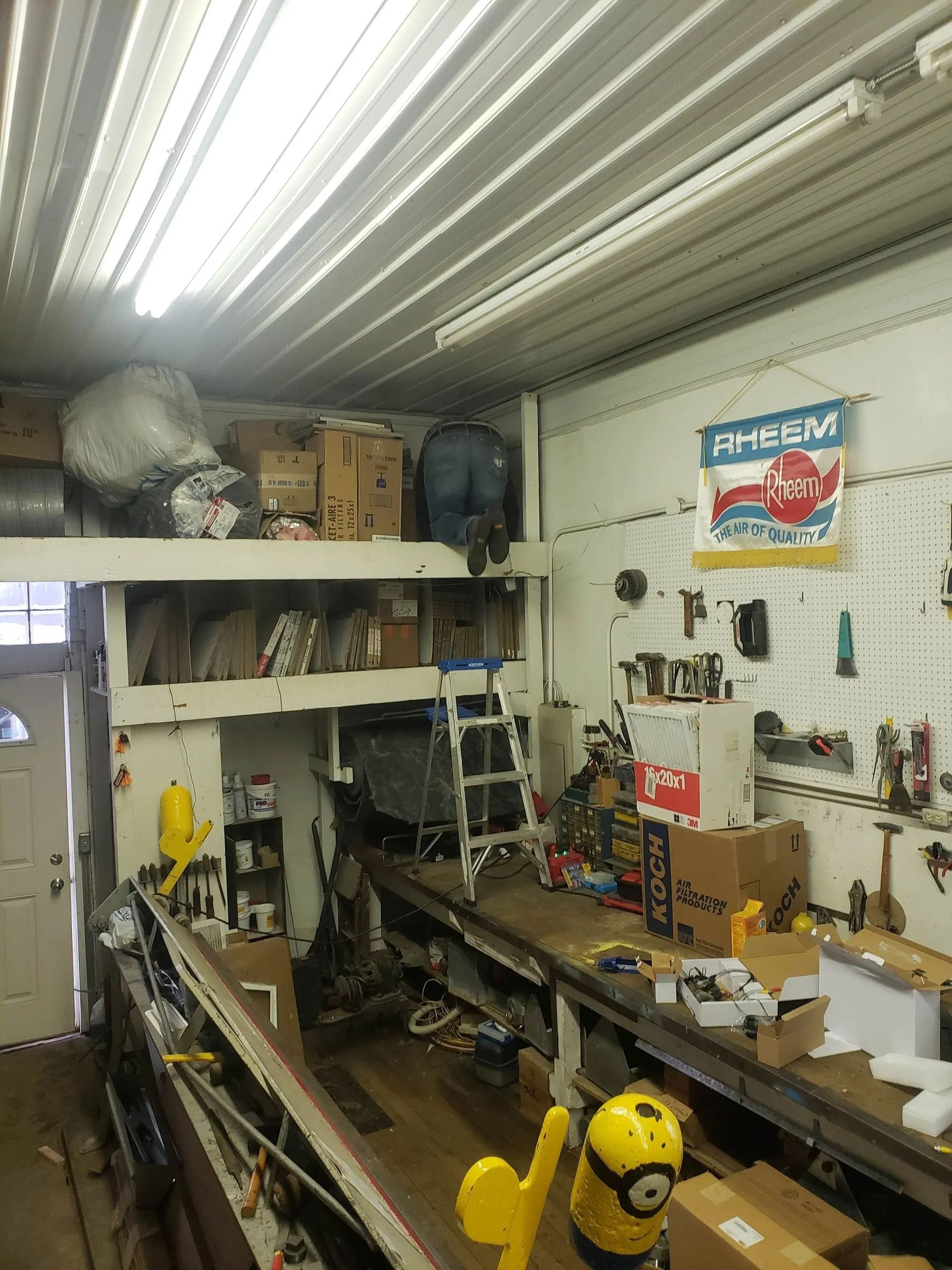 Cluttered workshop with shelves, tools, and a workbench under bright overhead lights.