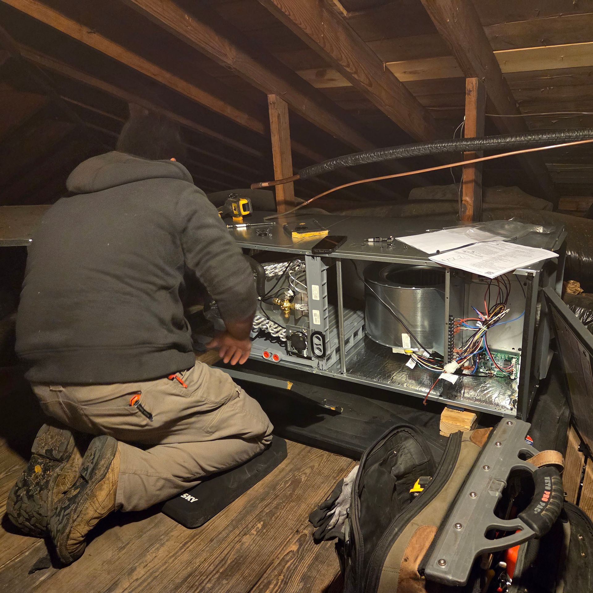 Person kneeling in an attic, working on an open electronic device with exposed wiring and tools.