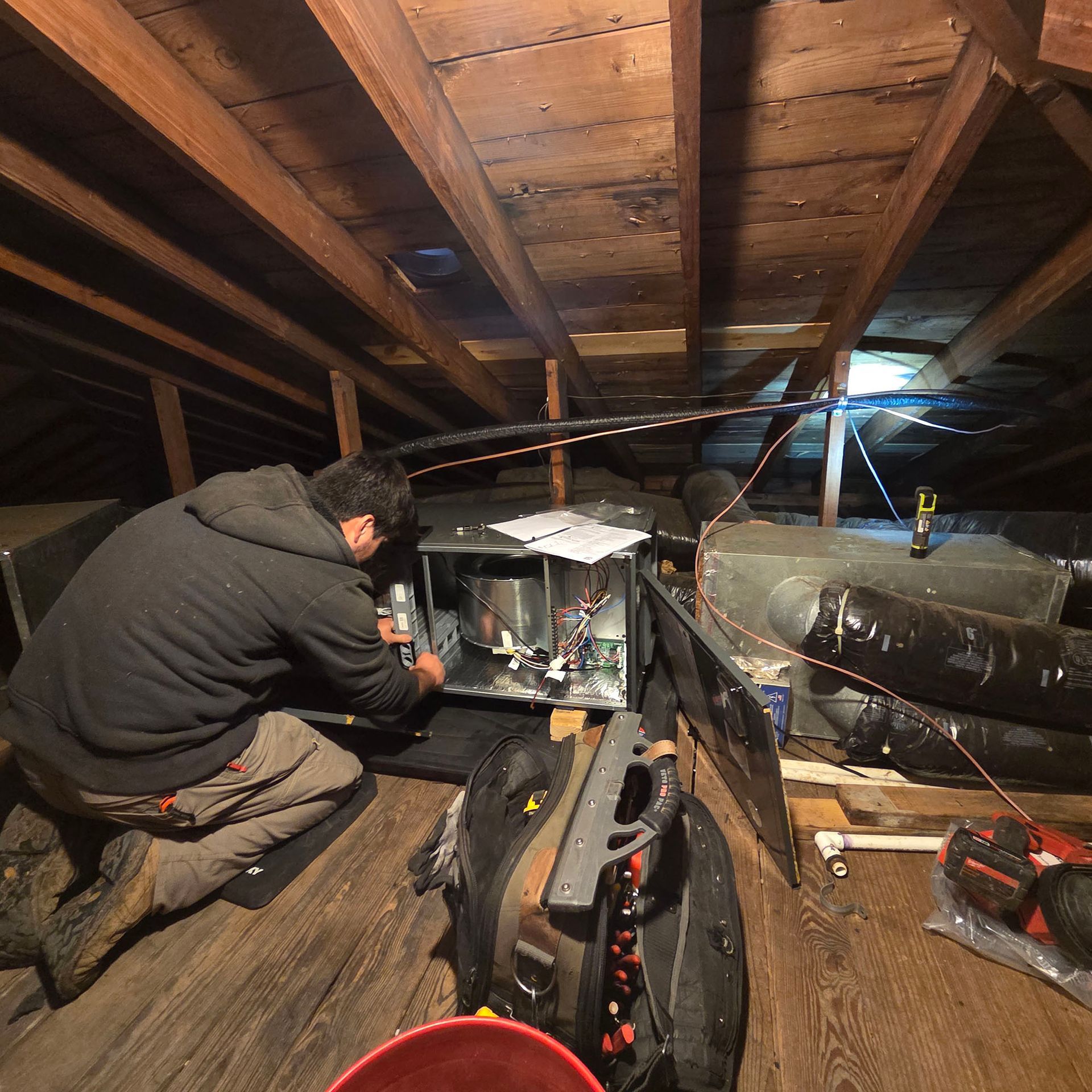 Person crouching in an attic, repairing HVAC equipment beside exposed ductwork and beams.