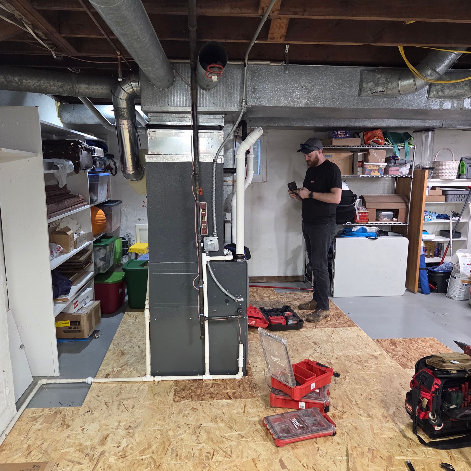Person working in a cluttered basement workshop beside a large furnace and exposed ductwork.