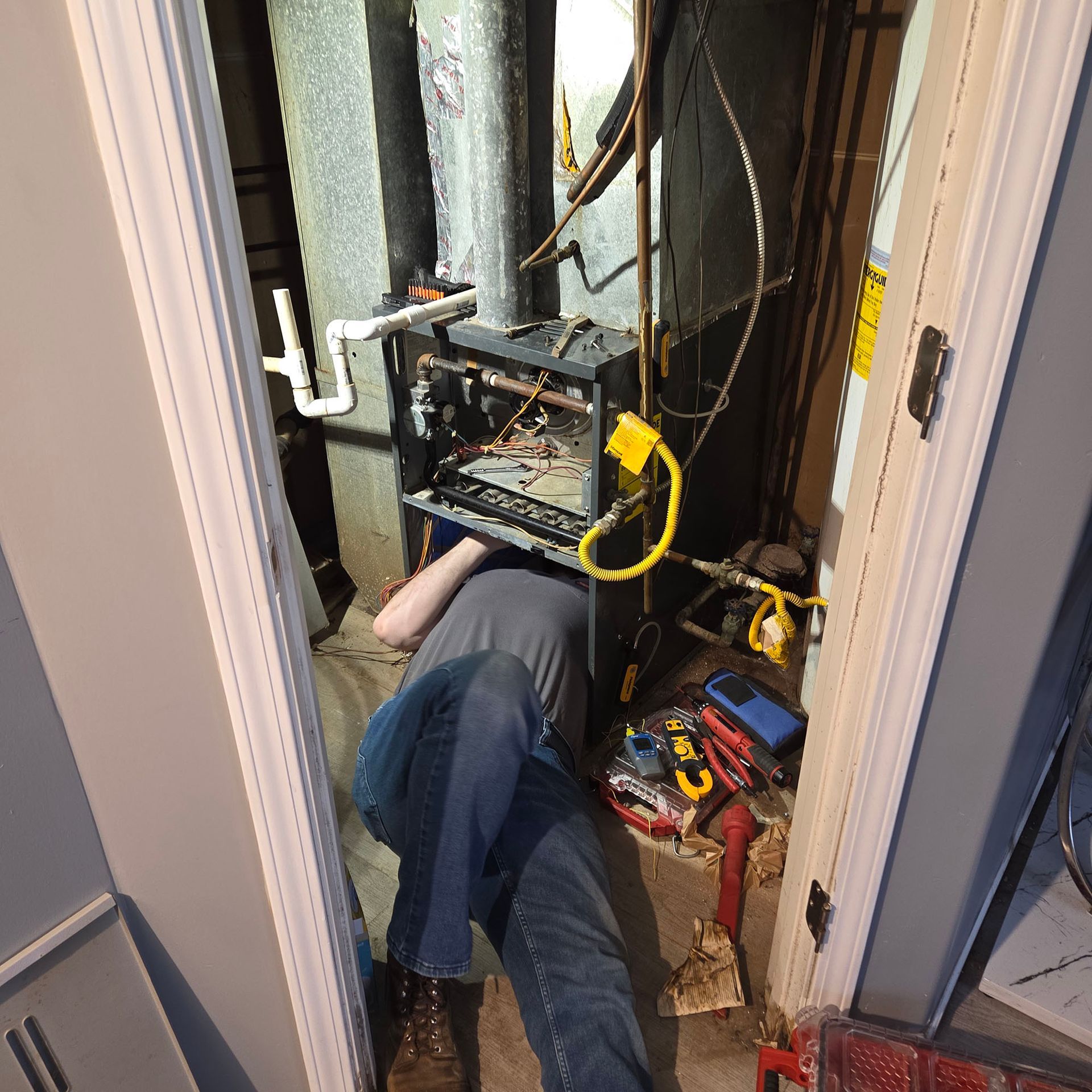 Technician lying in a narrow utility closet, working on exposed HVAC or electrical equipment with tools nearby