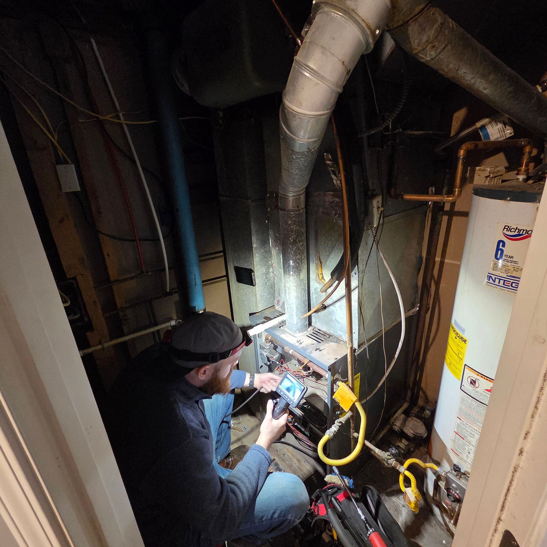 Technician inspecting wiring in a dim utility closet, with ductwork, pipes, and a water heater nearby