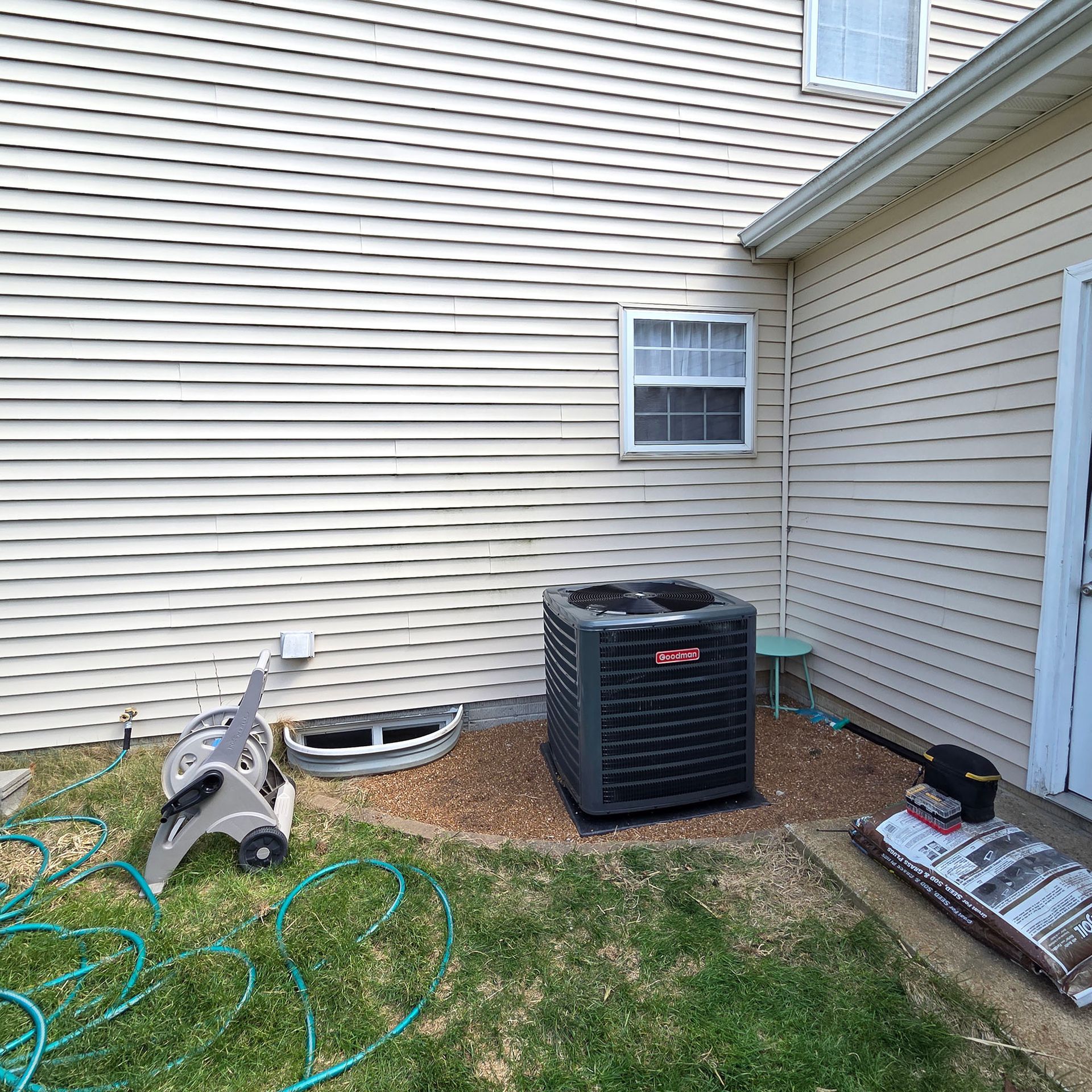 Backyard beside a beige house, with an air conditioner unit, hose, and yard tools on the grass.